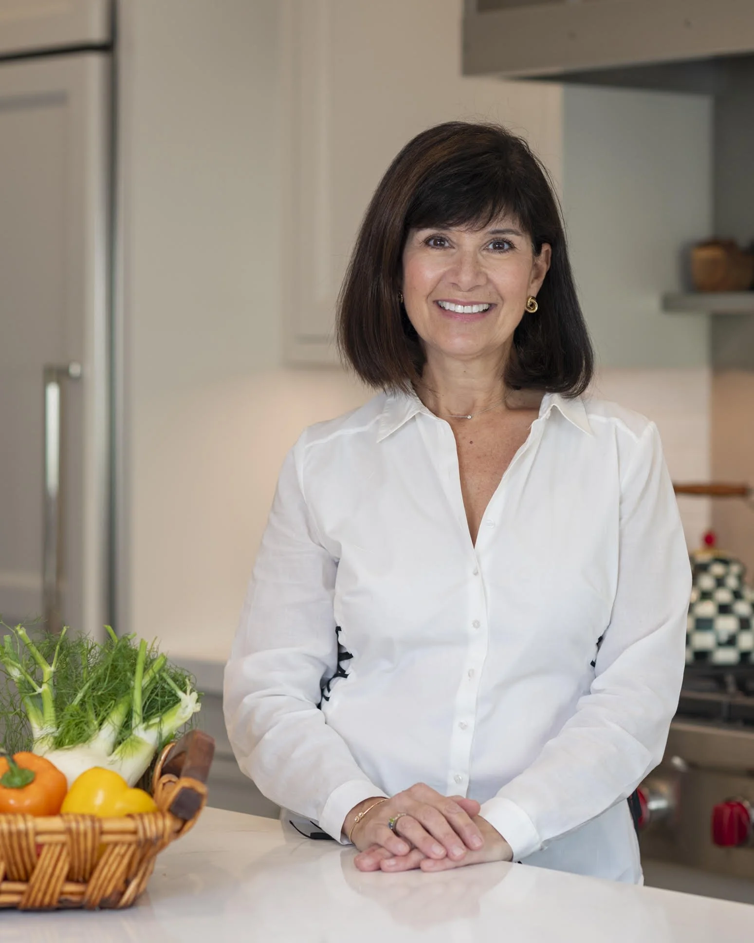 A woman with dark hair wearing a white shirt standing in a kitchen with a basket of vegetables on the counter.