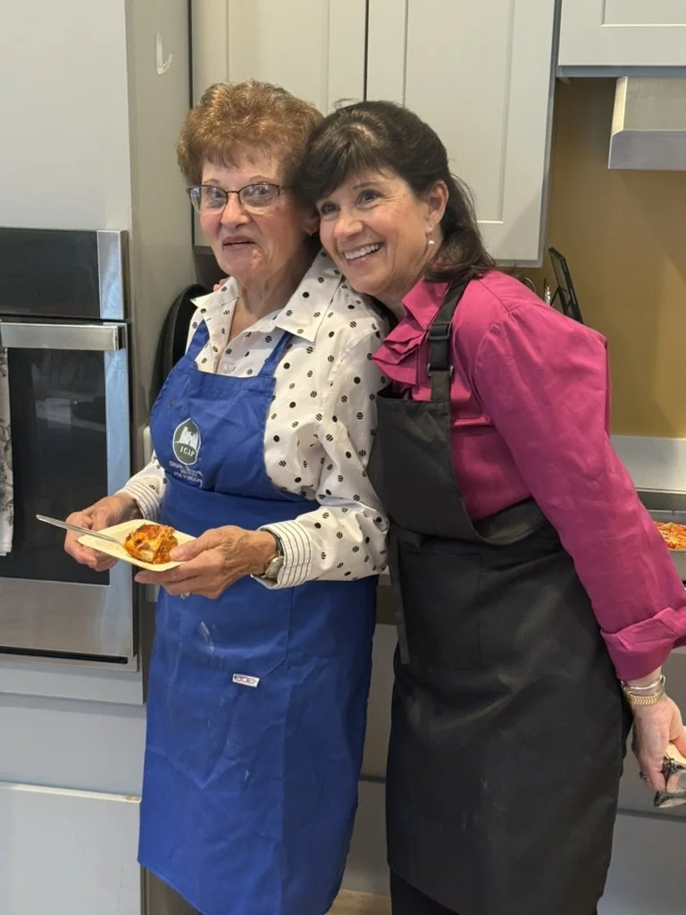 Two women are standing in a kitchen, smiling. The older woman on the left is wearing glasses, a white blouse with black polka dots, and a blue apron. She is holding a small plate with a slice of pizza. The younger woman on the right has dark hair and is wearing a pink shirt with a dark apron. They appear to be enjoying a moment together in a warm, homey kitchen environment.