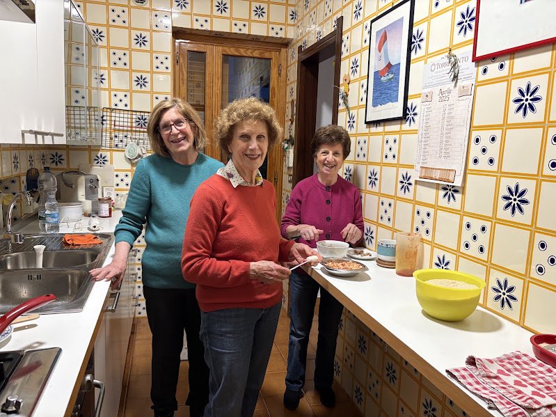 Three women standing in a kitchen, smiling, with the woman in the center holding a bowl and serving food, surrounded by kitchen utensils and colorful tile walls.