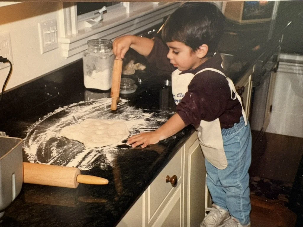 A young boy wearing a brown shirt, blue jeans, and an apron rolls out dough on a black kitchen countertop with a rolling pin nearby.