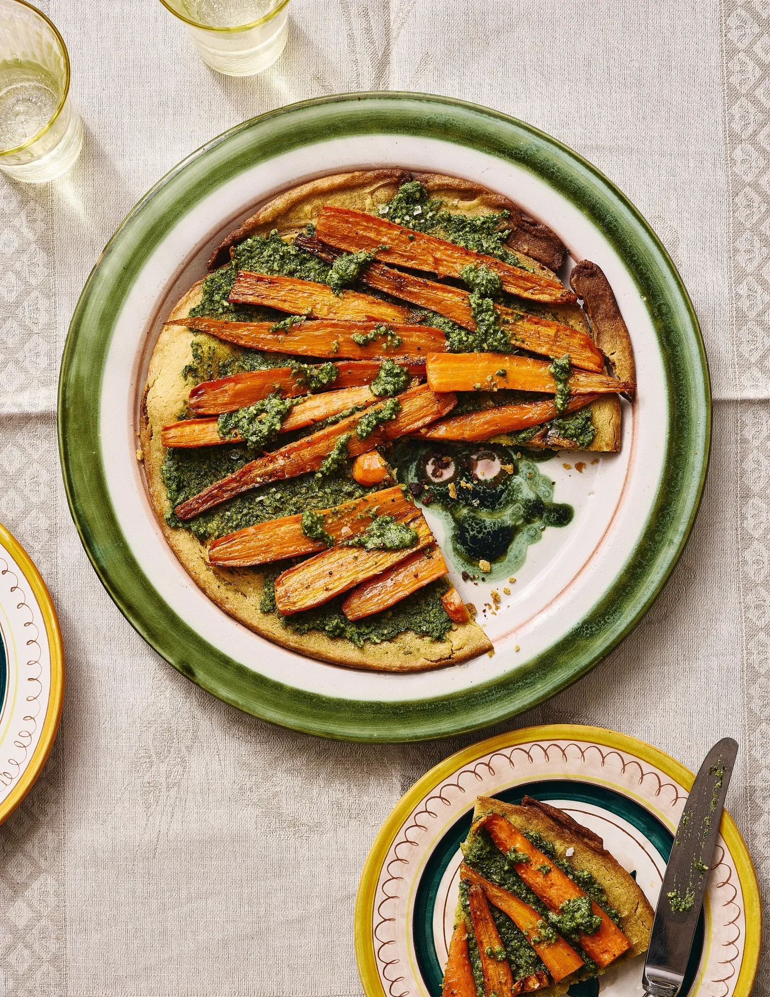 A green-rimmed plate with a slice of bread topped with roasted carrots and green herb sauce on a table set with matching dishware and glasses of water.