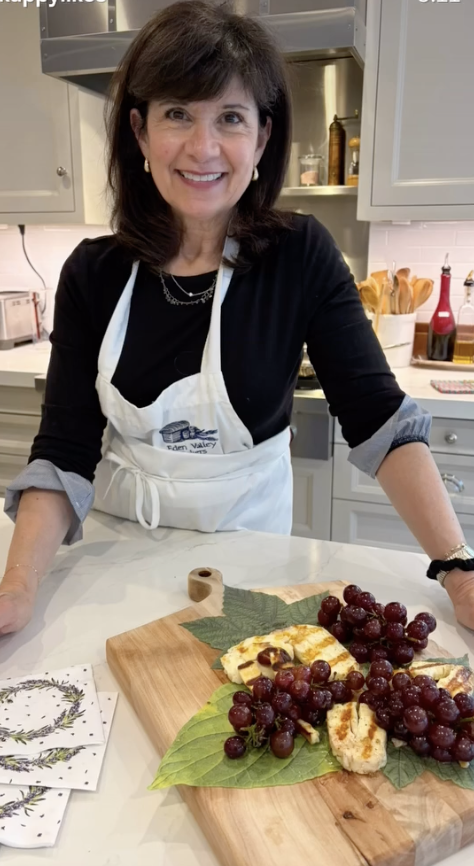 A smiling woman in a black top and white apron standing in a kitchen behind a cutting board with grapes and sliced cheese, with utensils and kitchen items in the background.