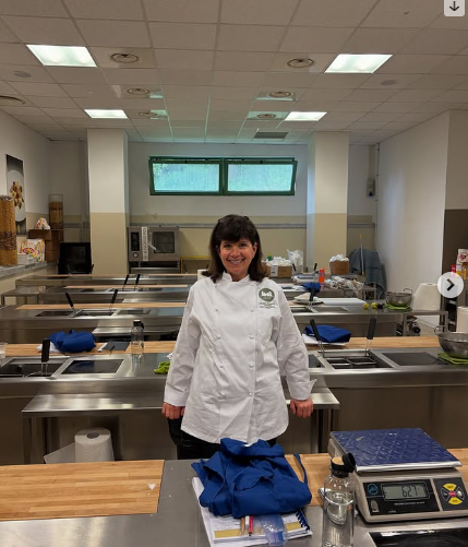 A woman in a white chef's coat smiling in a commercial kitchen with stainless steel counters and kitchen equipment.
