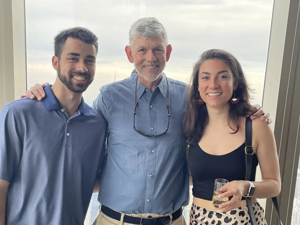Three people smiling and posing for a photo in front of a window with a view of a city skyline. The man in the middle has gray hair and a beard, wearing a light blue button-up shirt, and sunglasses hanging around his neck. The woman on the right has dark hair, wearing a black sleeveless top and leopard print pants, holding a glass of white wine. The man on the left has dark hair and a beard, wearing a blue polo shirt.