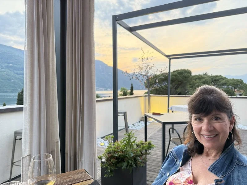 A woman smiling on a rooftop terrace during sunset with a lake and mountains in the background, surrounded by outdoor furniture and potted plants.