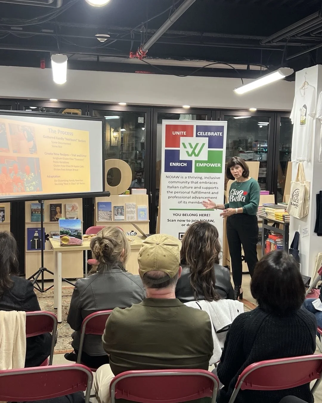 A woman giving a presentation to a seated audience in a bookstore or community space. Behind her are bookshelves and a colorful sign with the words 'Unite', 'Celebrate', 'Enrich', and 'Empower'. A large screen displays a slide with the title 'The Process' and information about recipes and adaptations. The audience is paying attention, and some are wearing casual clothing.