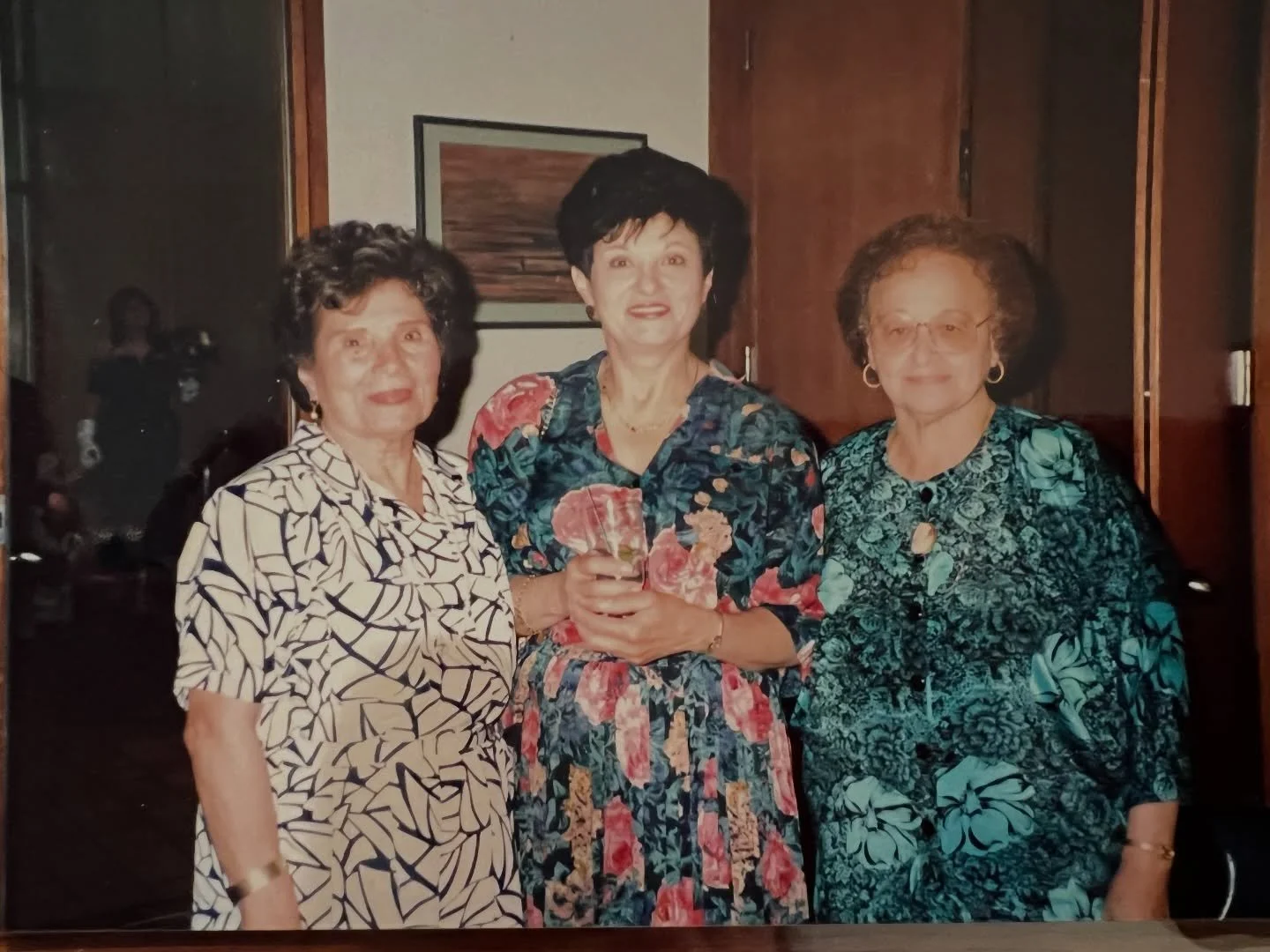 Three women standing together indoors, dressed in floral and patterned dresses, smiling at the camera.