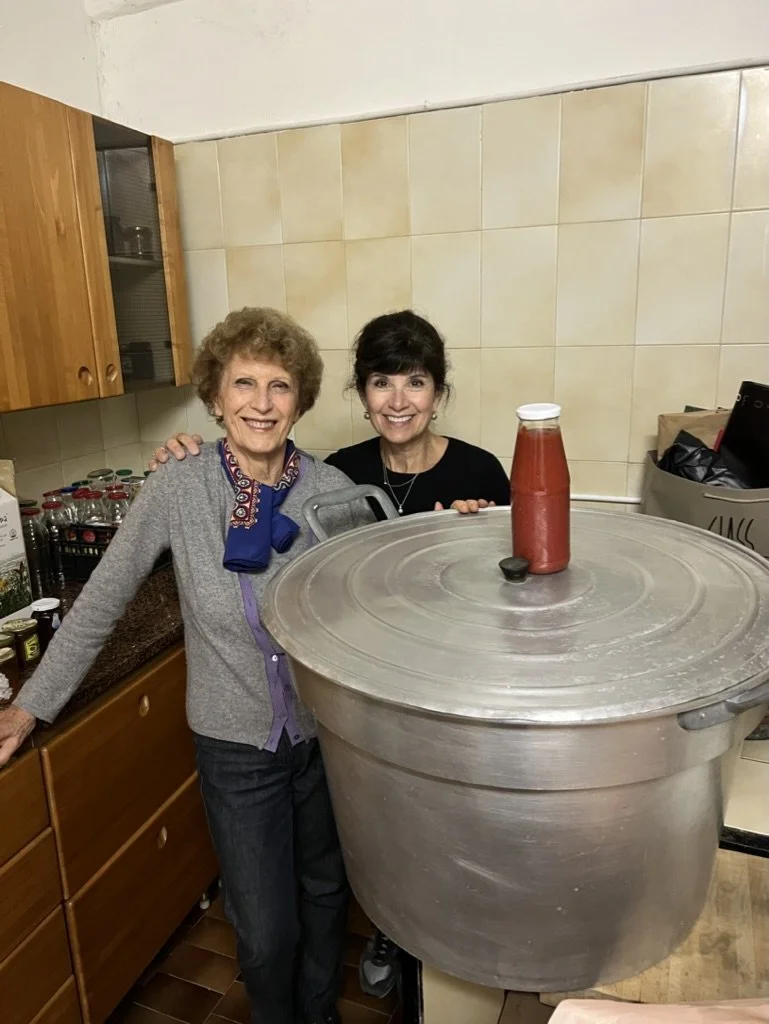Two women standing together in a kitchen, smiling, behind a large steel cooking pot with a bottle of ketchup on top.