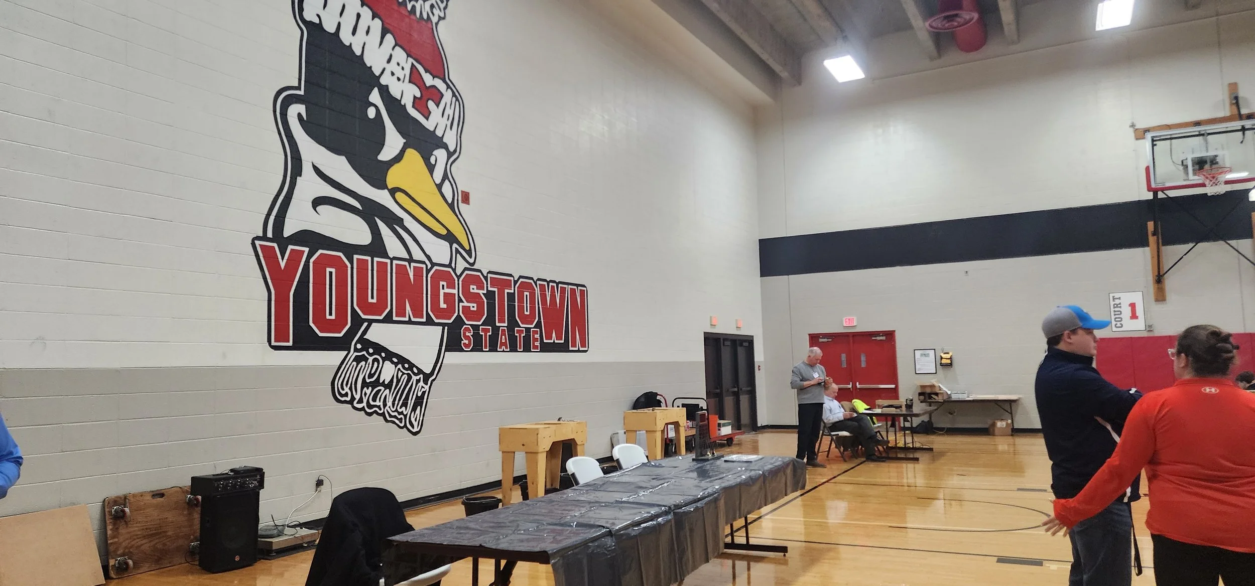 Indoor gymnasium with a large wall decal reading 'Youngstown State' featuring a stylized bird logo. Several people are present, some standing and others sitting. Tables are set up against the wall, and basketball backboards are visible.