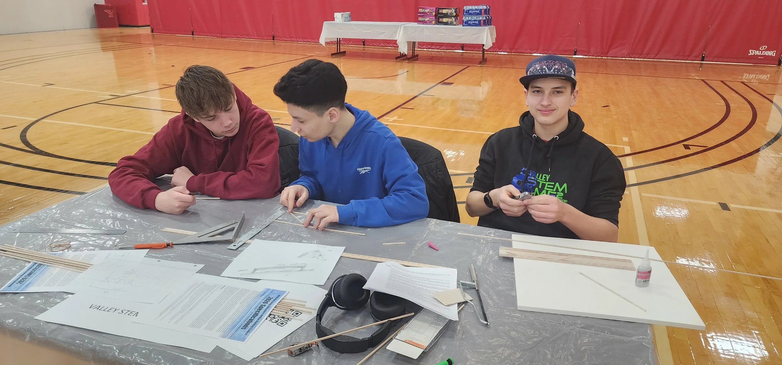 Three young men sitting at a table in a gymnasium, working on a STEM project with wood and tools.
