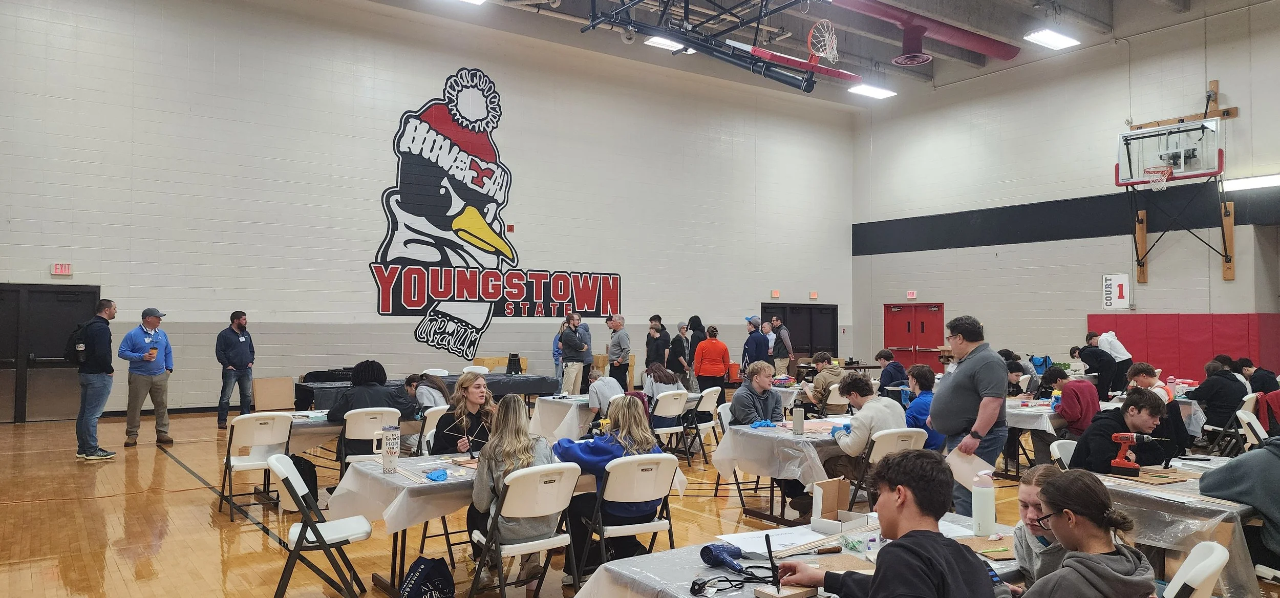 A gymnasium filled with people sitting at tables and standing, some engaging in activities or conversations, with a large YOUNGSTOWN STATE logo featuring a penguin mascot on the wall. There are basketball hoops and a sign indicating Court 1.