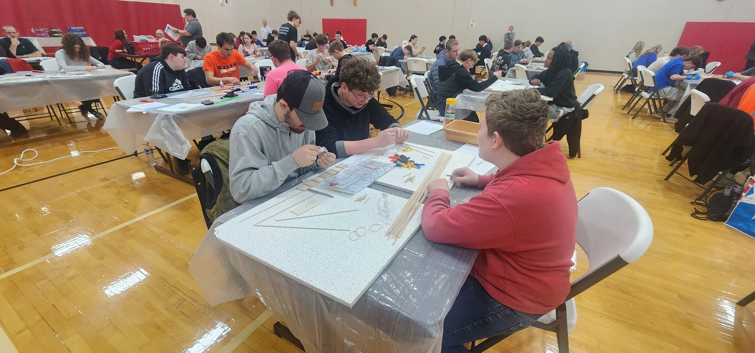 Group of young people sitting at long tables in a gymnasium, engaged in tabletop activities, with materials like rulers, sticks, and papers, in a room with red walls and wooden floor.