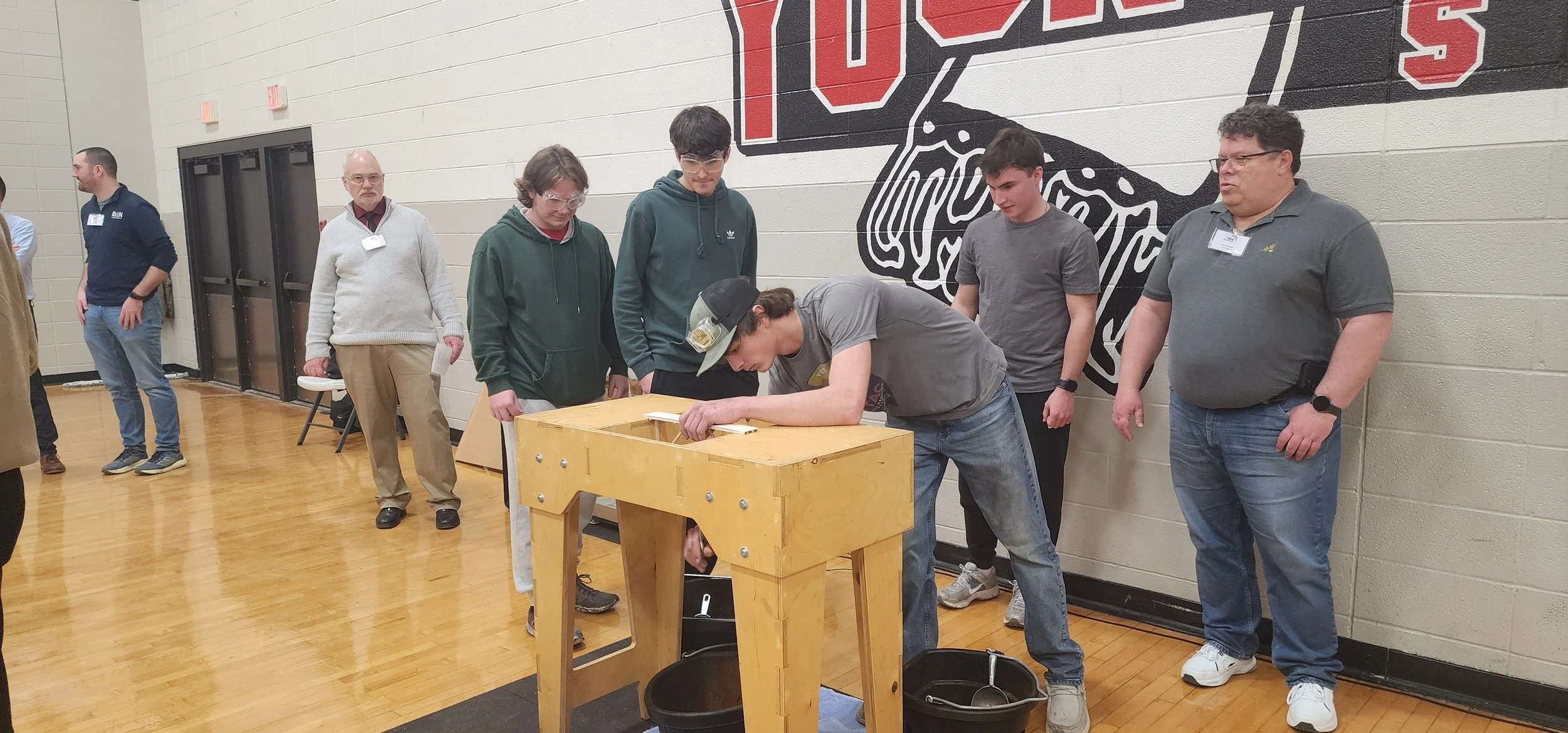 A group of people standing and watching a person working on a woodworking project at a wooden table in a gymnasium or indoor space with a mural on the wall.