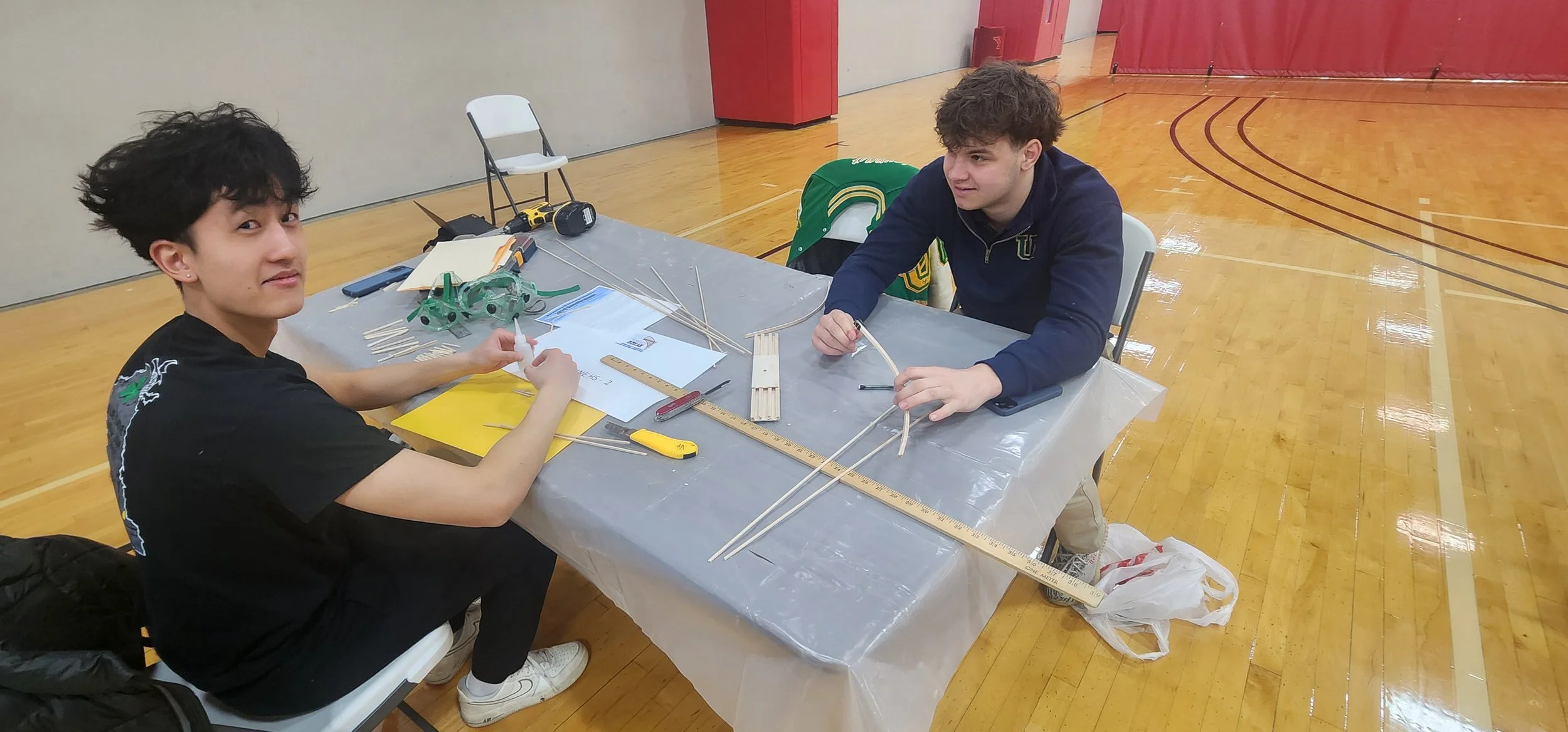 Two young men working on a robotics project at a table in a gymnasium.