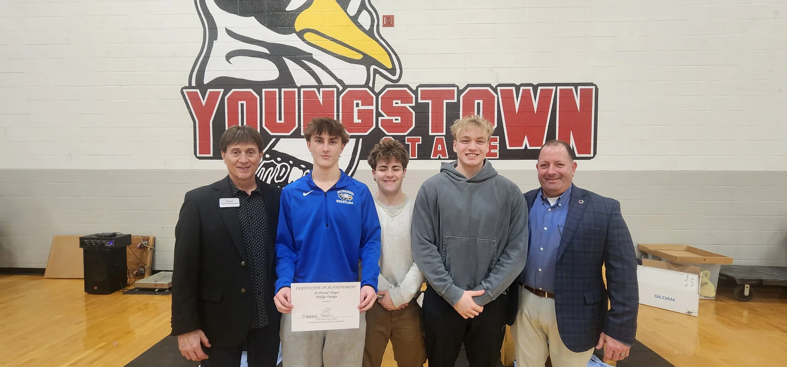 Four young men and two adult men standing together in front of a large logo that reads 'YOUNGSTOWN TAE' on a gymnasium wall, with one of the young men holding a certificate of achievement.