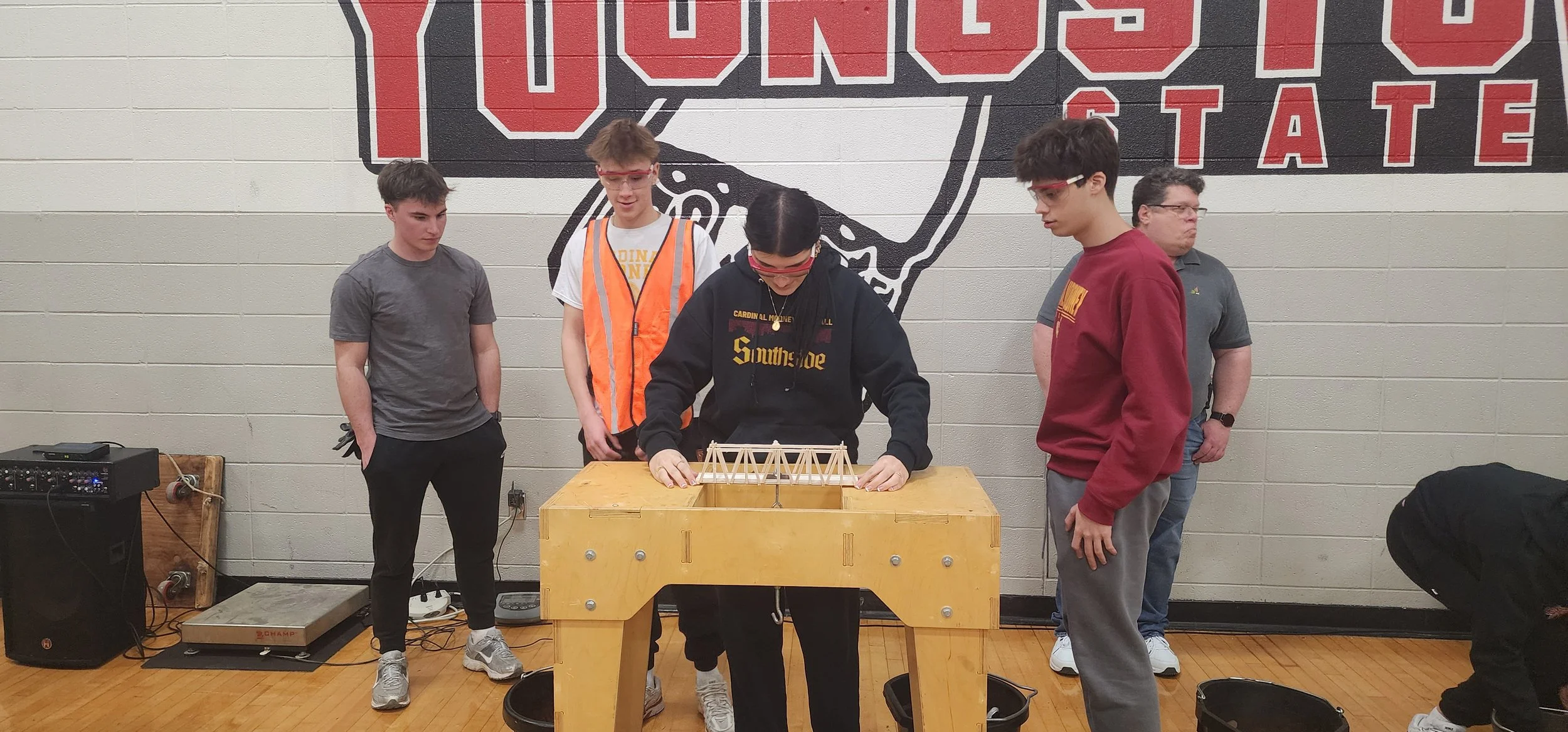 Group of five teenagers working on a bridge model at a school event, with a large Ohio State sign painted on the wall behind them.