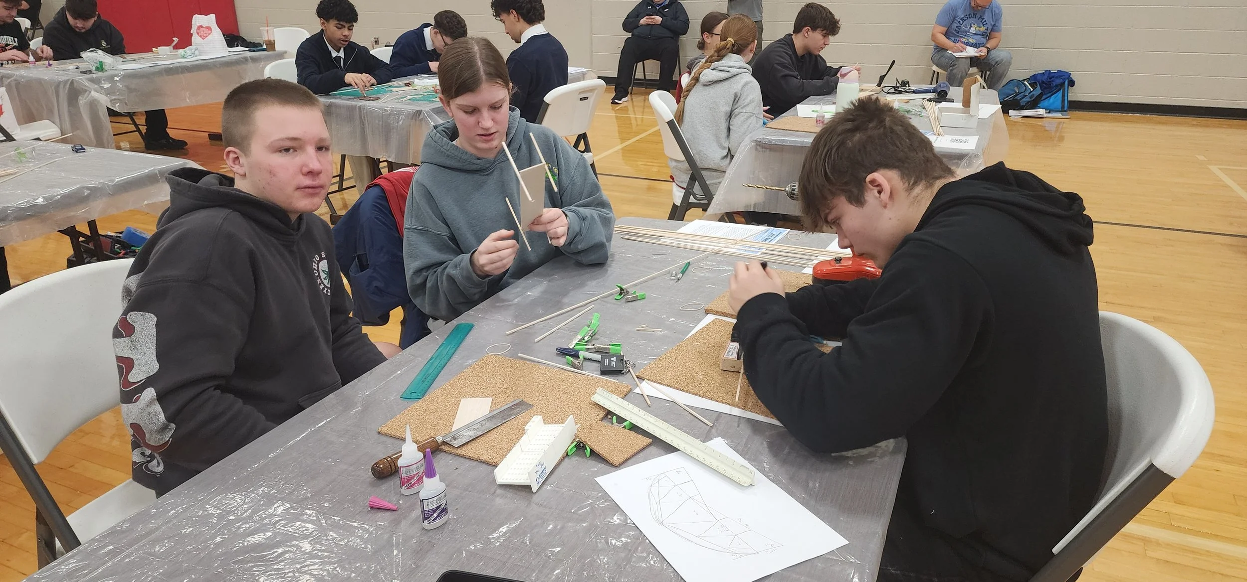 Several students are working on a STEM project at a table in a school gymnasium. They are using wooden sticks and measurement tools, with some students focused on their work and others looking at the camera.