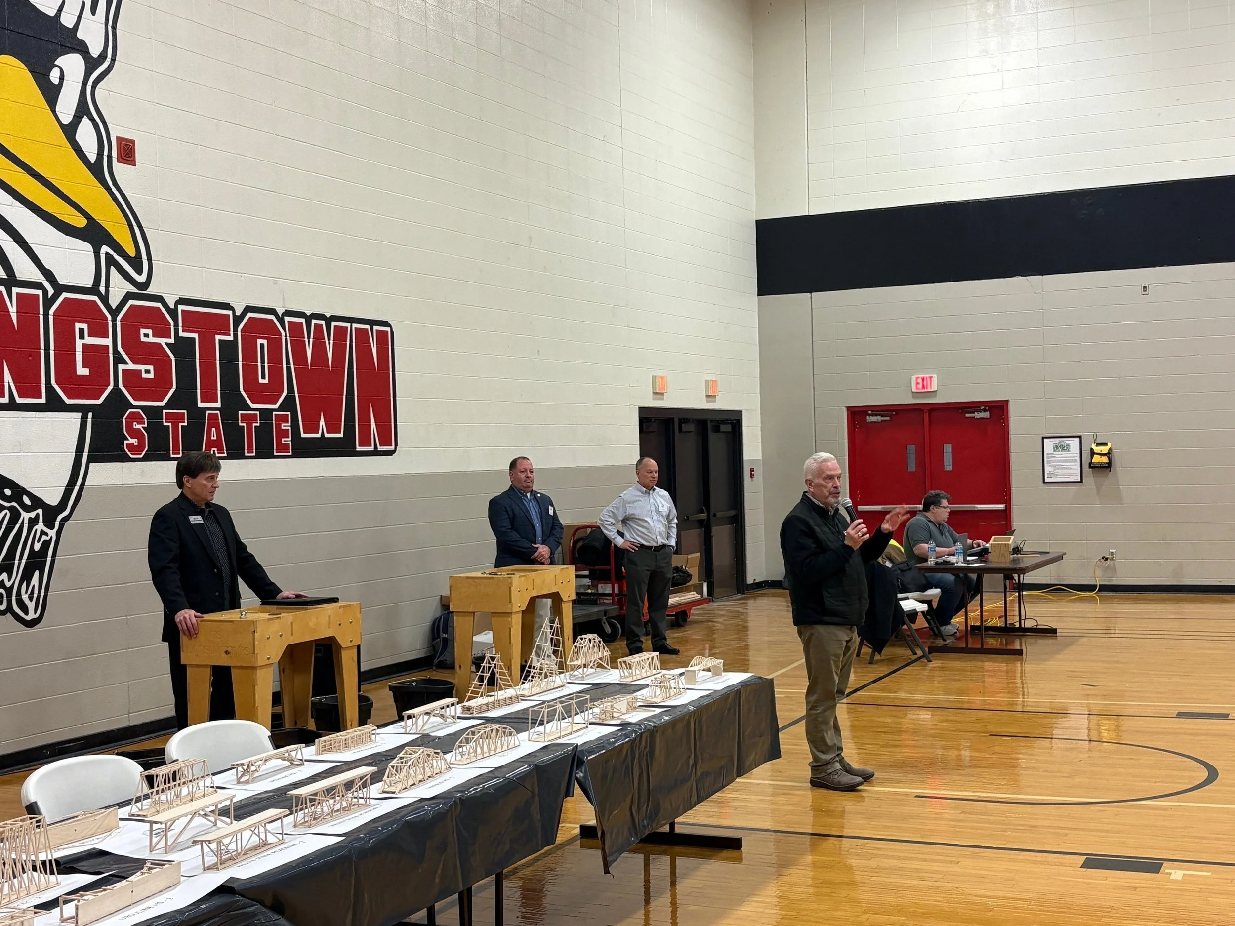 People standing at a table with architectural models in a gymnasium with a large wall featuring a logo of a hawk and the words 'GSTOWN STATE' on the wall.