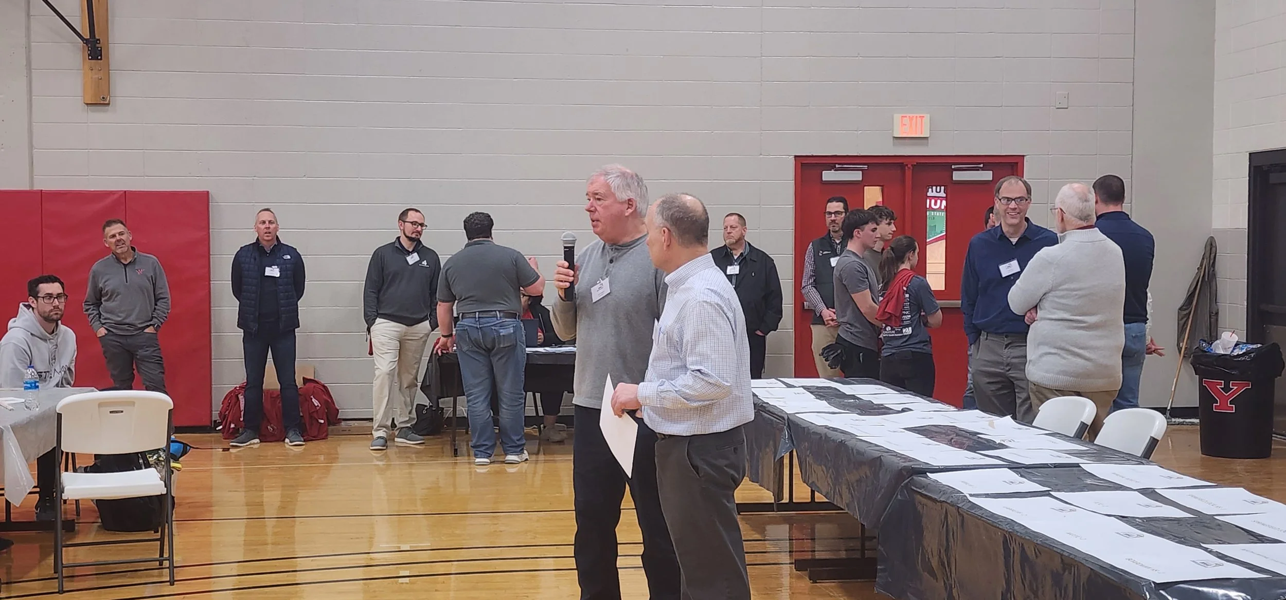 A group of people gathered in a gymnasium, some standing and some sitting, engaging in conversations and looking at displays or tables with printed materials. One man is speaking into a microphone, and others are wearing conference badges.