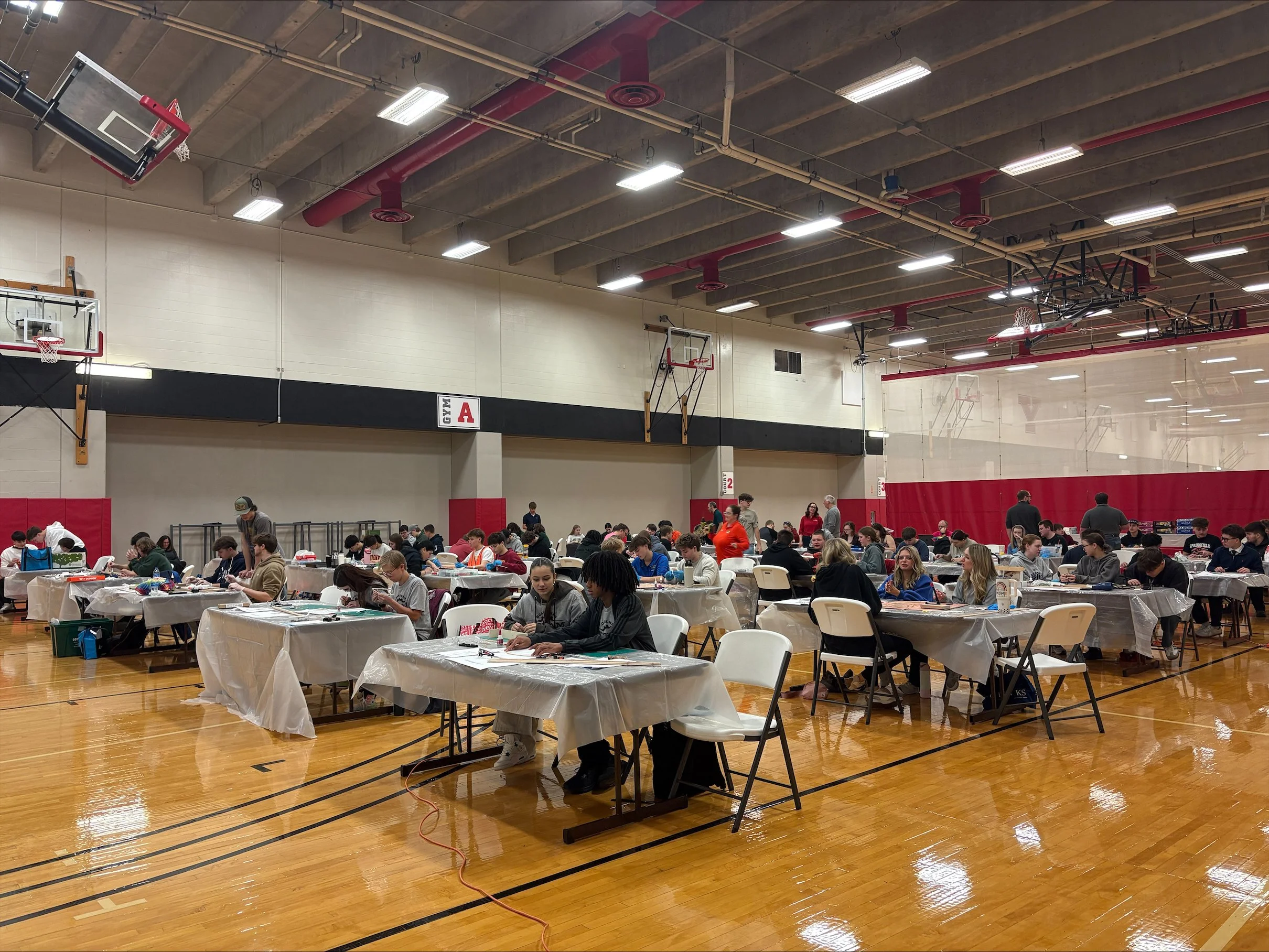 A gymnasium filled with people participating in a craft or activity event, with tables covered in plastic tablecloths and various supplies, basketball hoops, and a high ceiling with red and beige piping.