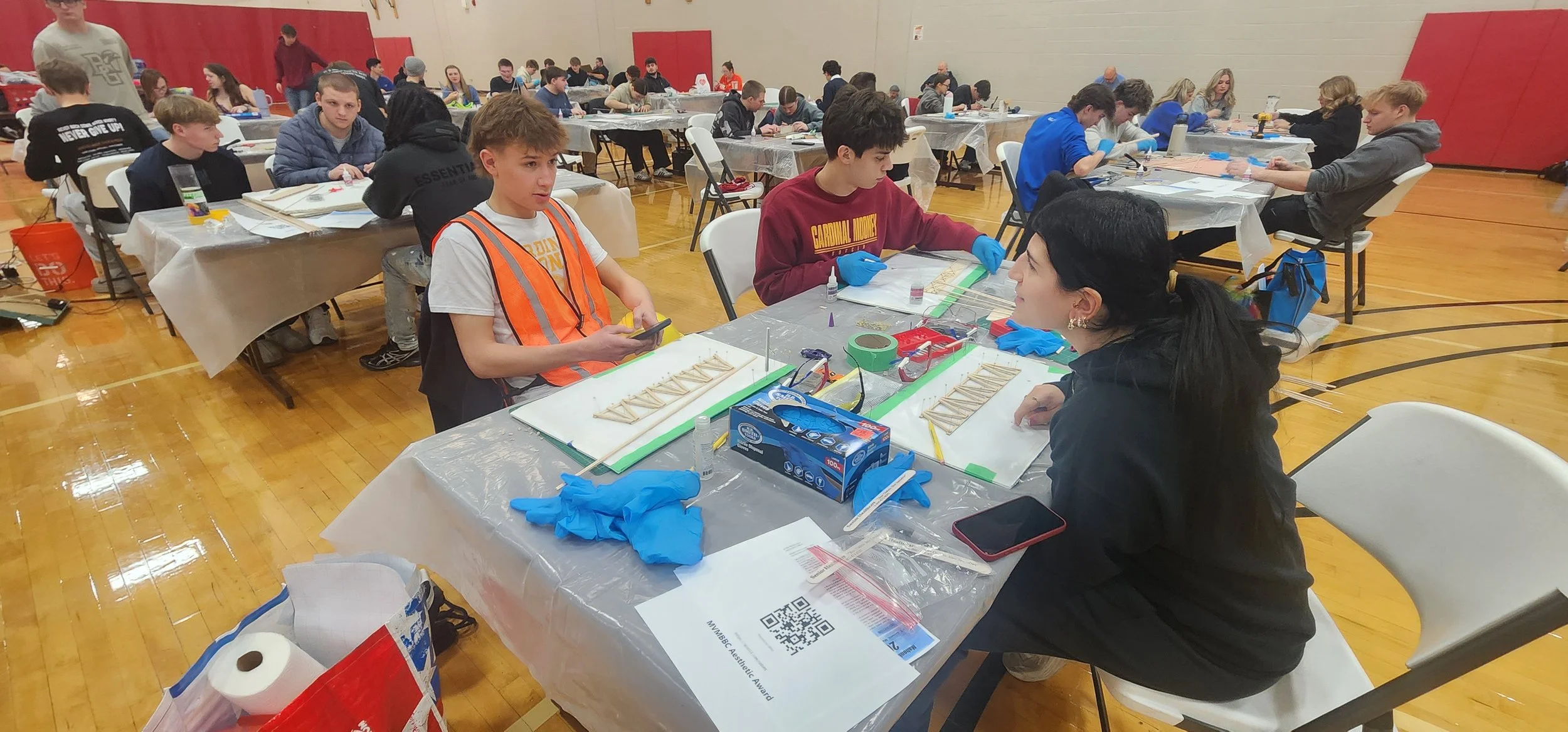Students and instructor at an indoor STEM competition working on bridge-building projects with supplies and tablets, multiple tables and participants visible.