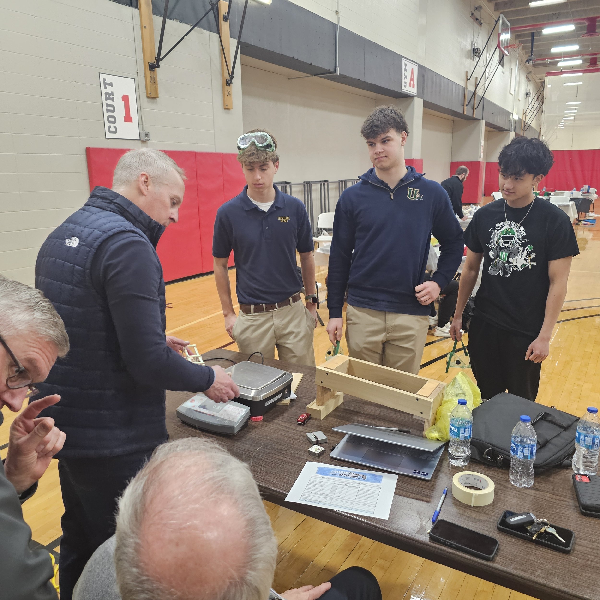 A group of young men standing around a table in a gymnasium, one man appears to be demonstrating or explaining something on a laptop. The table has various items such as water bottles, a tape roll, and electronic devices. There are tables and chairs 