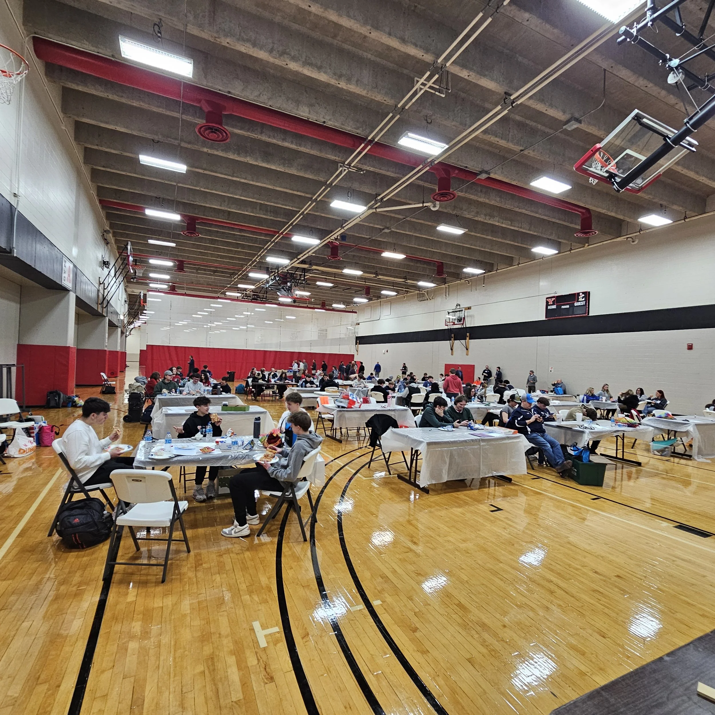 Indoor gymnasium filled with students sitting at tables and eating, with a basketball hoop and scoreboard in the background.