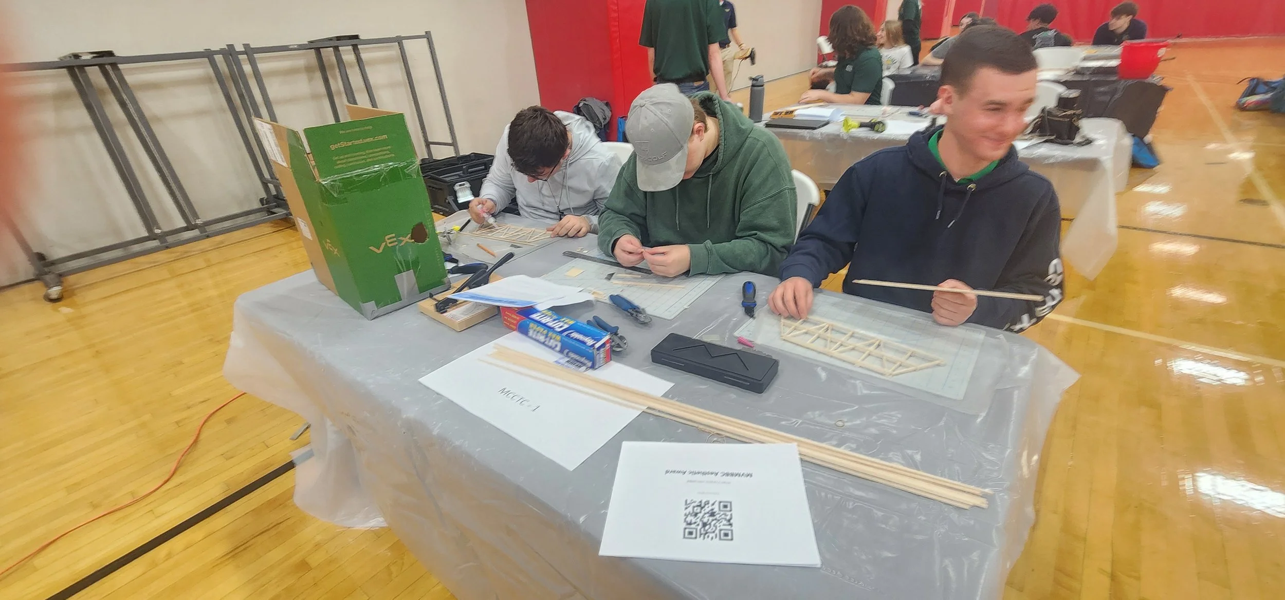 Several students working on model building projects at a table in a gymnasium during a STEM event. There are tools, model parts, and a QR code on the table.