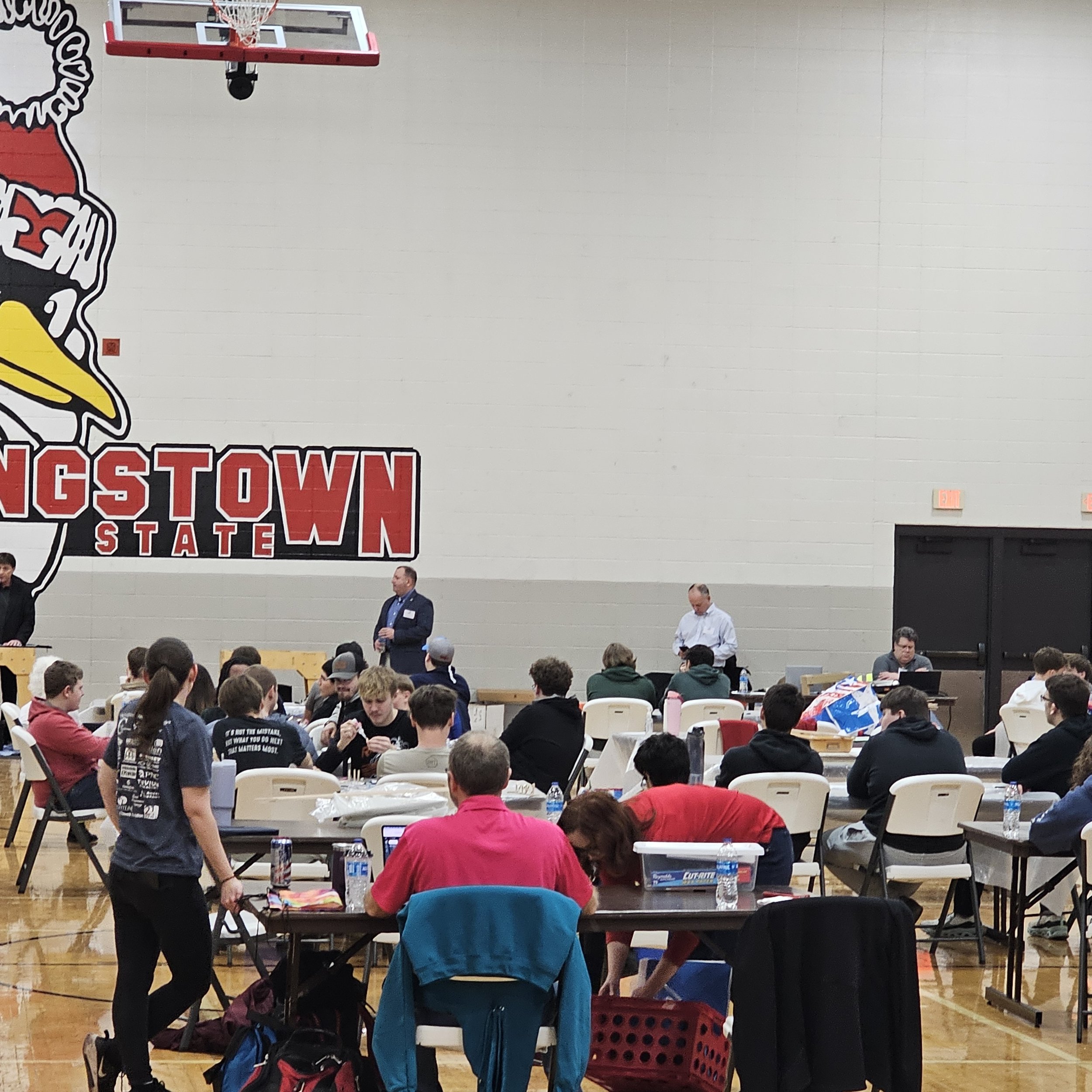 Inside a gymnasium with people attending an event, a large wall display reads 'Gaston State' with a logo of a cardinal bird. People are seated at tables with water bottles and bags.
