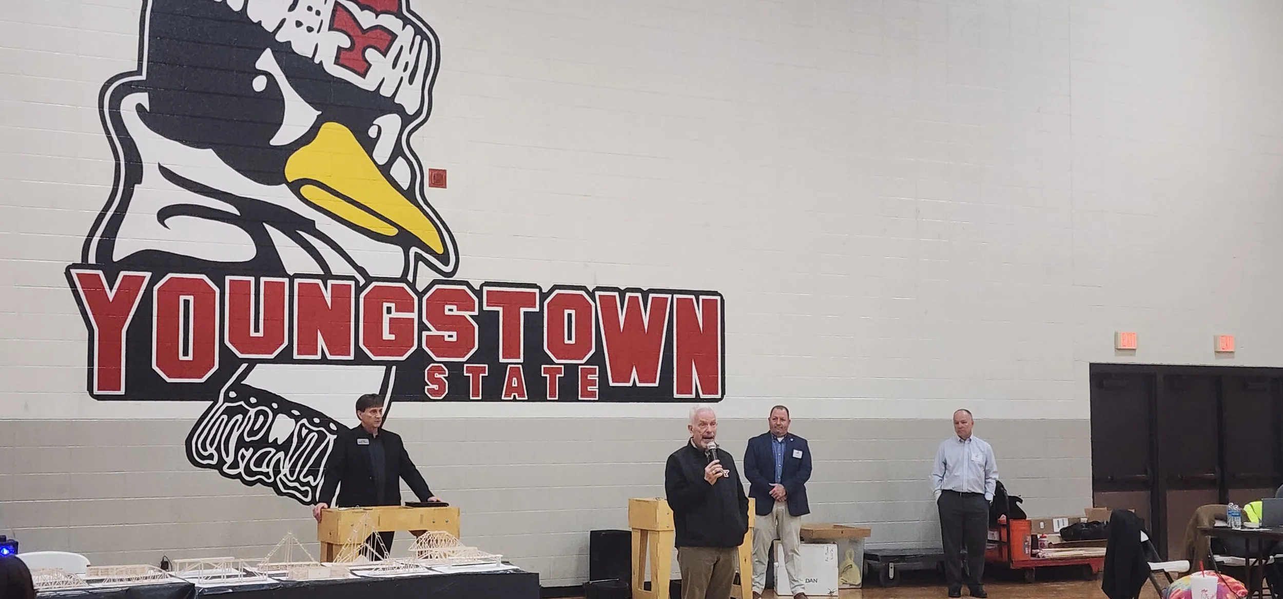 A group of men standing in front of a large wall graphic of a bird with a yellow beak and the words 'YUNGSTOWN STATE' on a sports team or school logo. One man is speaking into a microphone, while others look on.