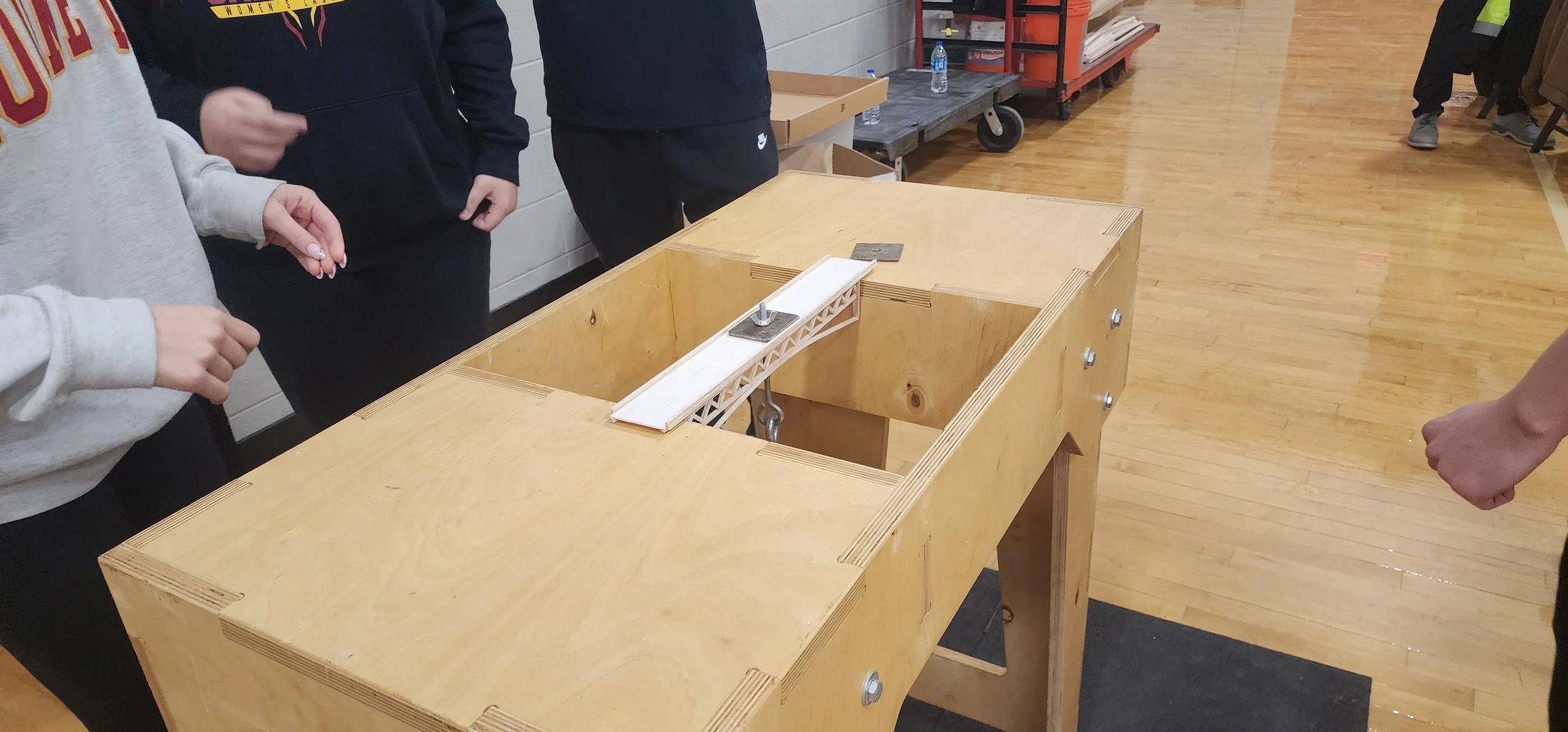 People standing around a wooden table with a small bridge model on it, inside a gymnasium or indoor sports facility.
