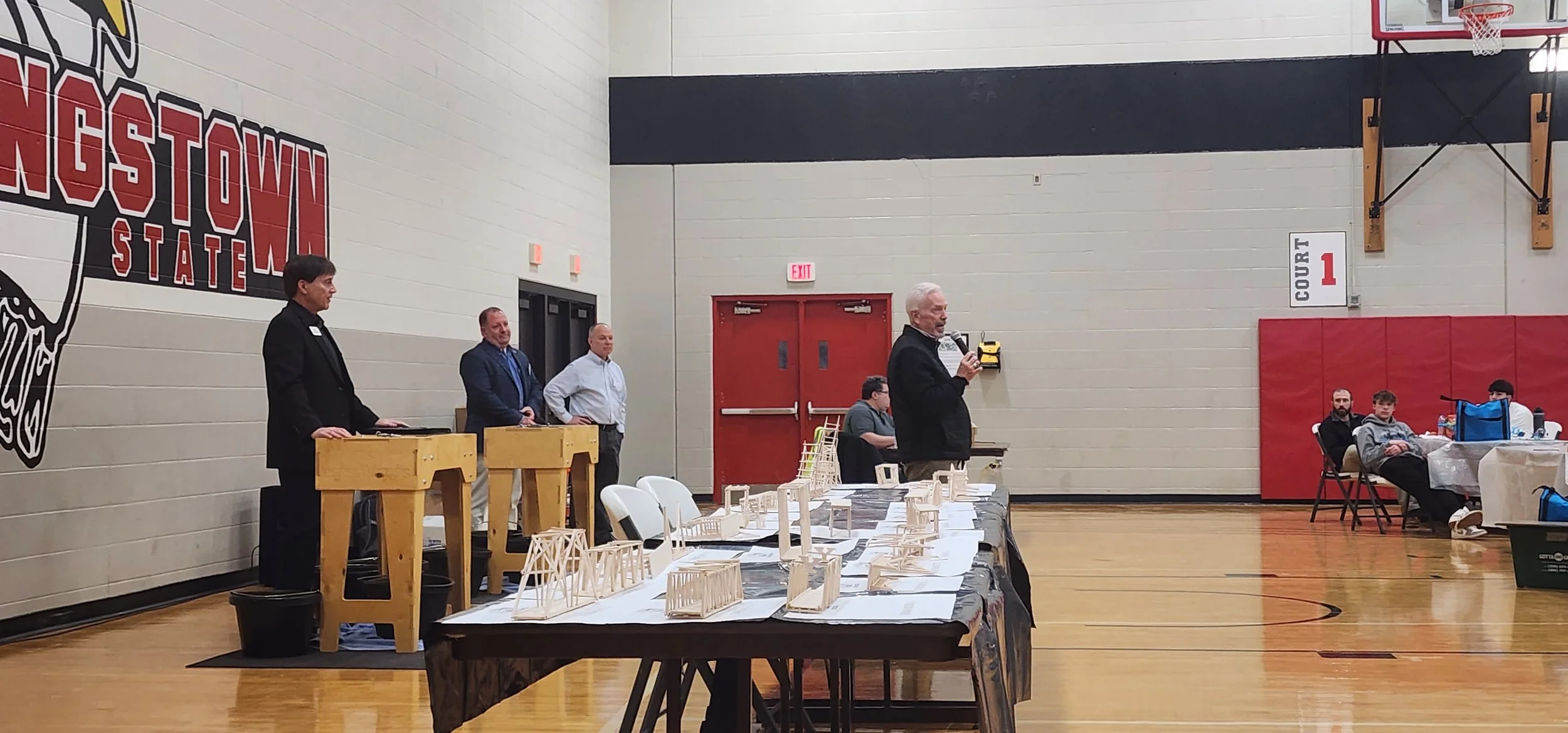People attending an indoor presentation or auction in a gymnasium, with tables displaying architectural models and a speaker speaking into a microphone.