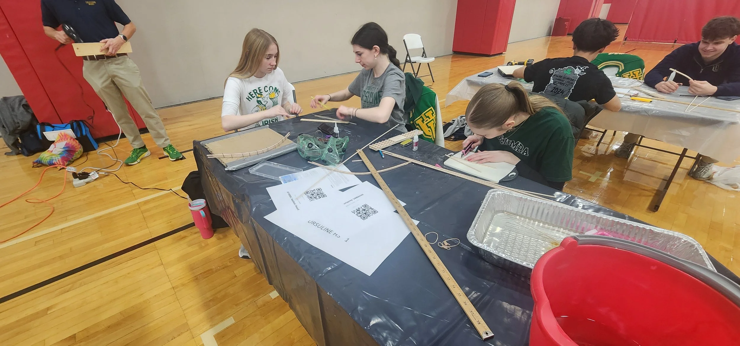 Group of students working on projects at tables in a gymnasium, with tools, rulers, and materials scattered on the tables.