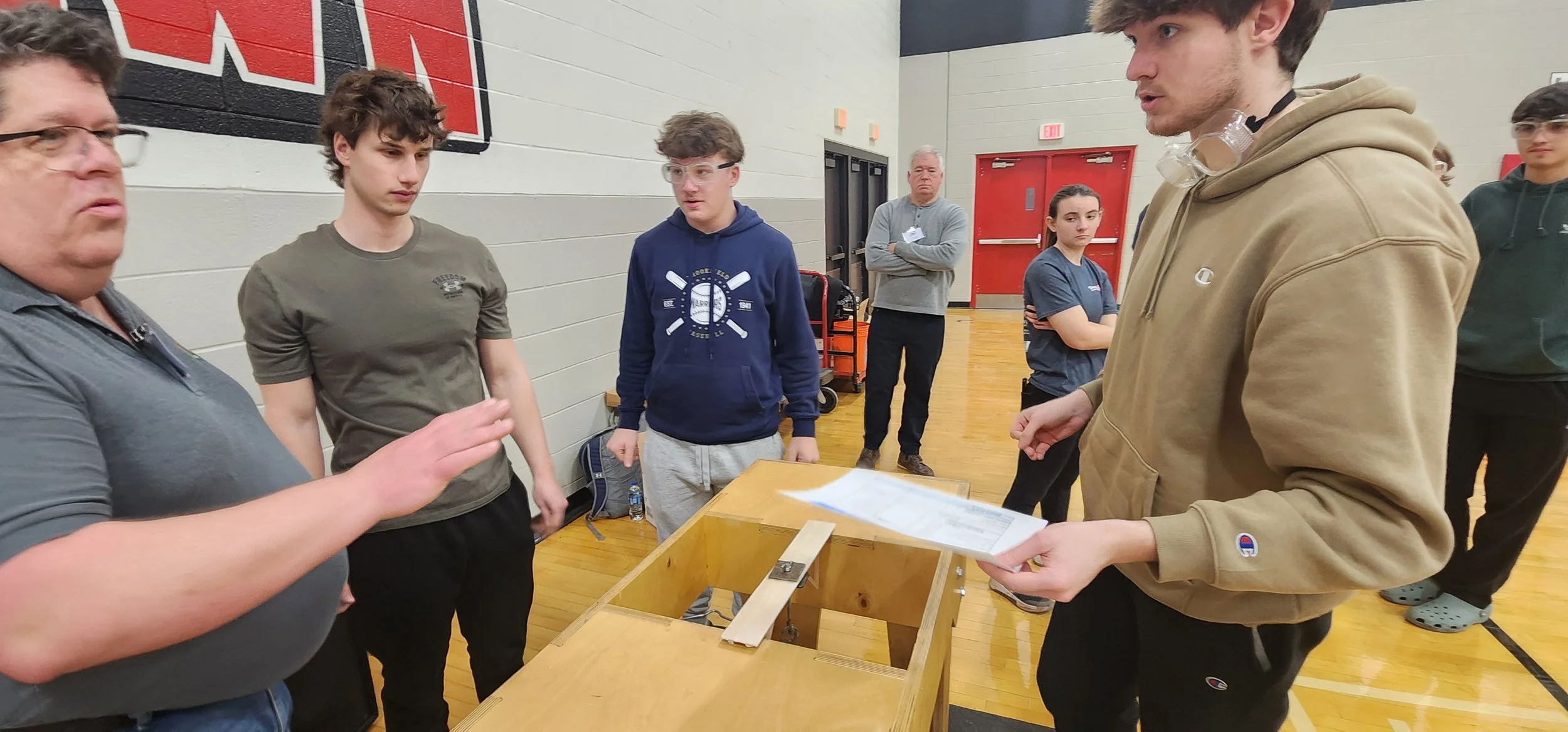 Group of people in a gymnasium engaged in a discussion, with a man in a gray shirt on the left speaking and a young man in a beige hoodie holding papers on the right.