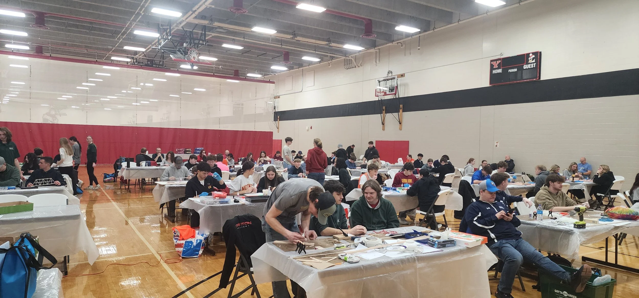 People working at tables in a gymnasium, engaging in a hands-on activity, likely a workshop or event, with a basketball hoop and scoreboard visible on the wall.