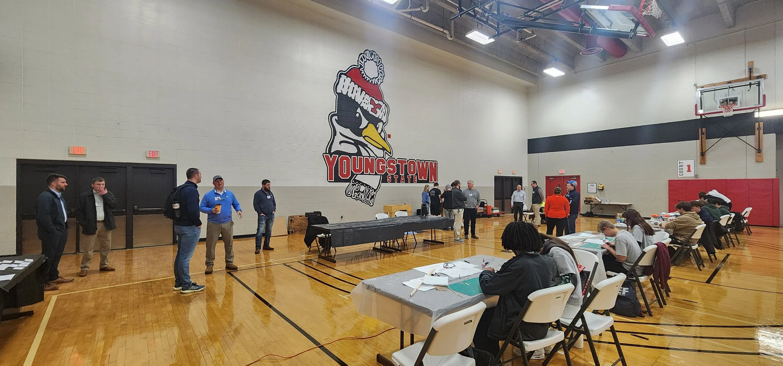 People gathered in a gymnasium for a meeting or event, with a large mural of a bird wearing a winter hat and the words "Youngstown State" on the wall.