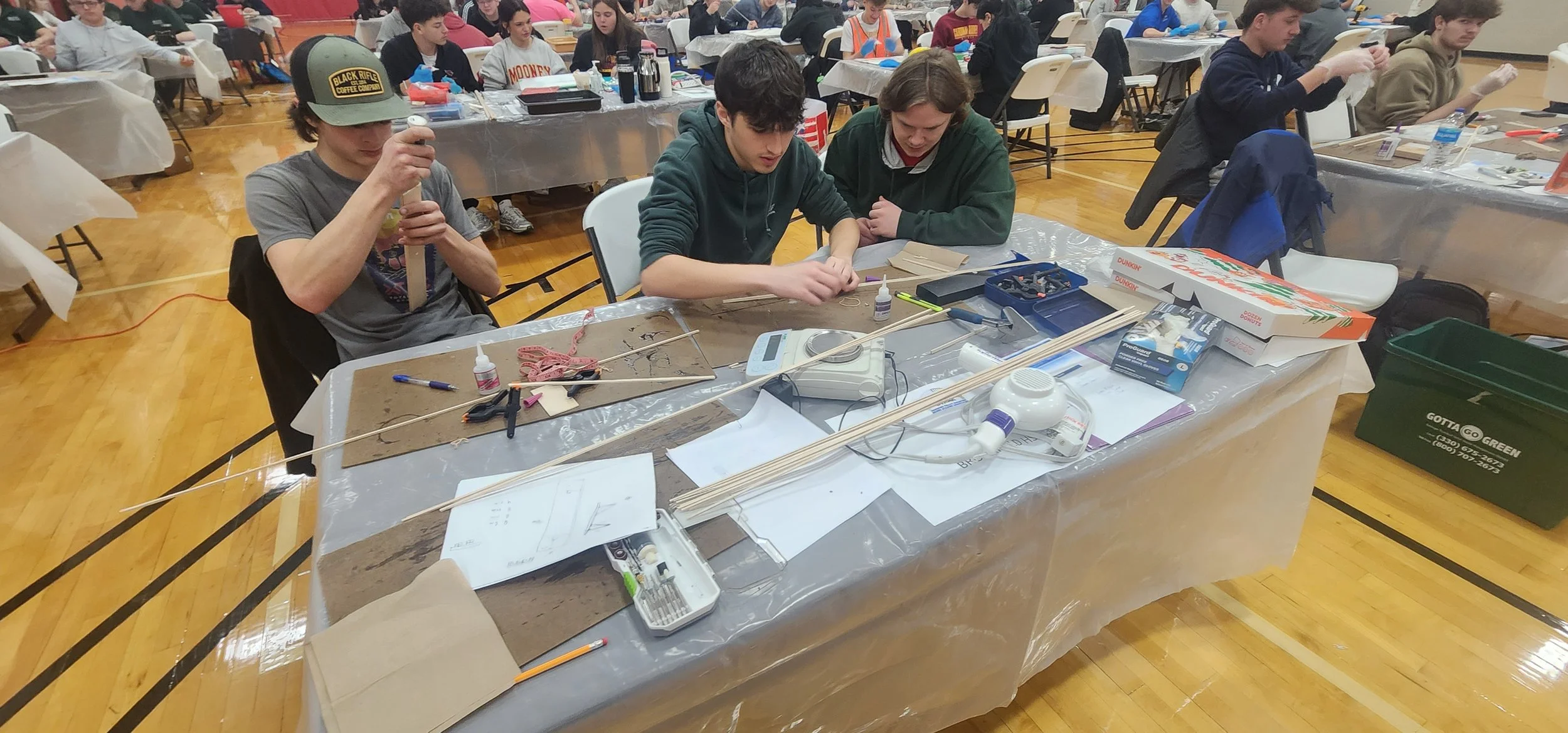 A group of young people working on a project at a long table covered with tools, wood pieces, glue, and papers in a gymnasium or community center setting.