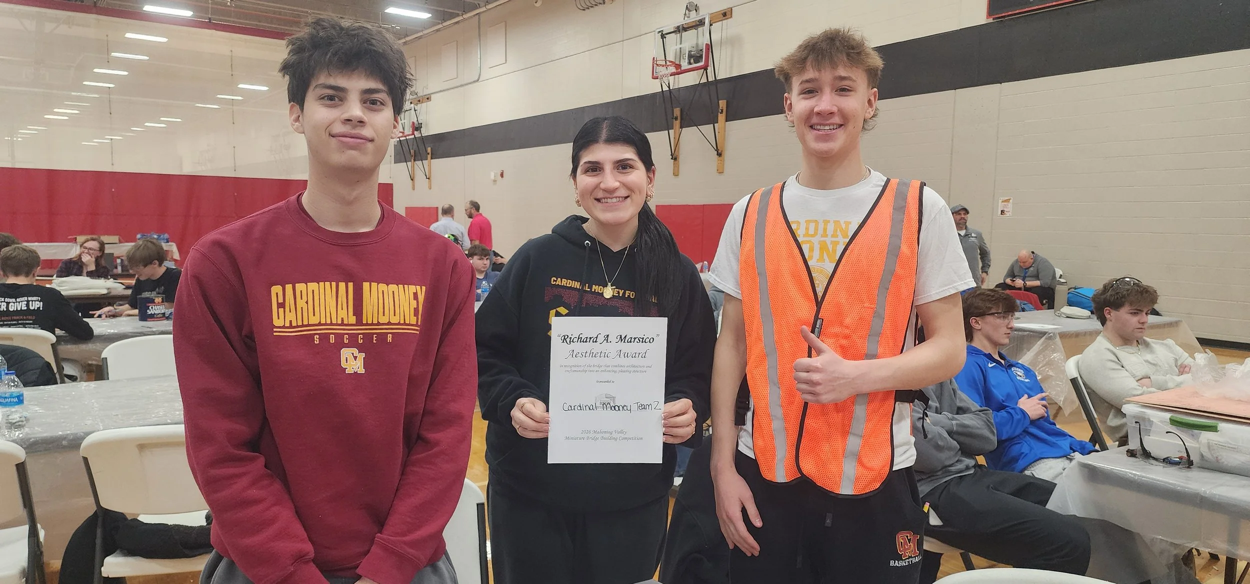 Three teenagers standing together in a gymnasium, with one girl holding an award certificate. The girl in the middle wears a black hoodie with a gold medal around her neck. The boy on the left wears a maroon sweatshirt, and the boy on the right wears