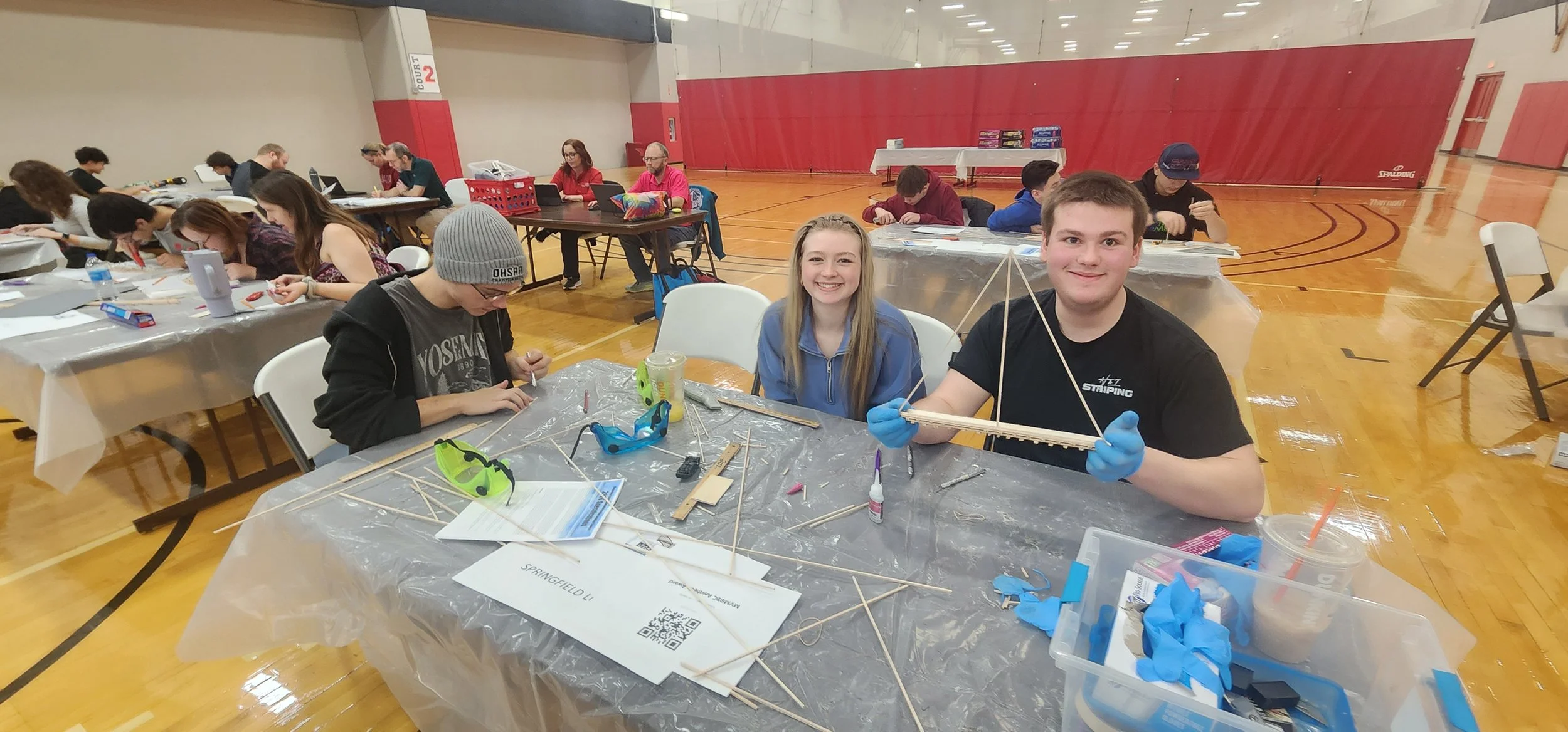 A group of children and teenagers at a table in a gymnasium working on a STEM project with wood sticks, glue, and tools. Two teenagers in the foreground are smiling, one holding a wooden structure.