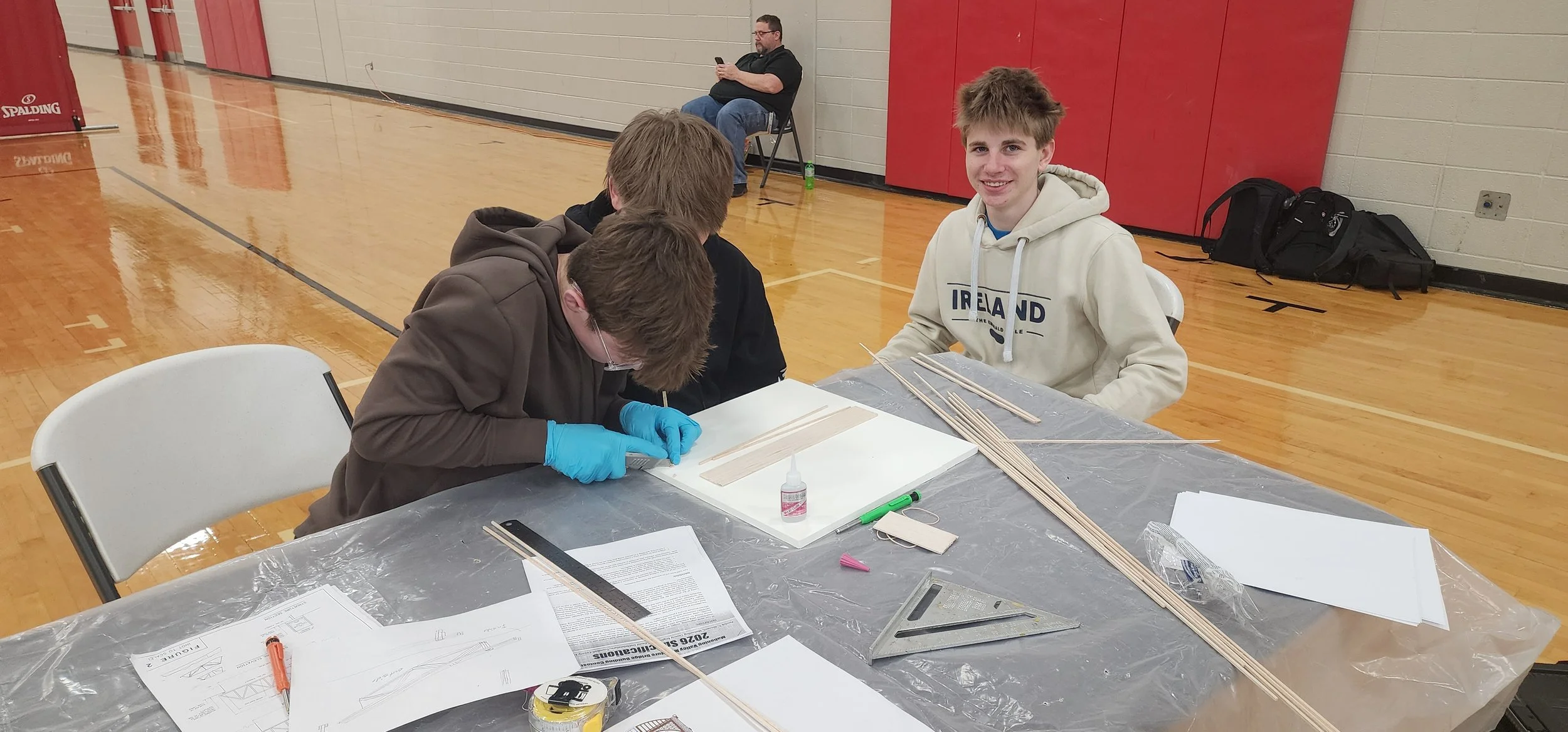 A group of young men sitting at a table in a gymnasium. One young man is working on a project with wooden sticks, while another is smiling at the camera. An older man is sitting in the background, looking at his phone.