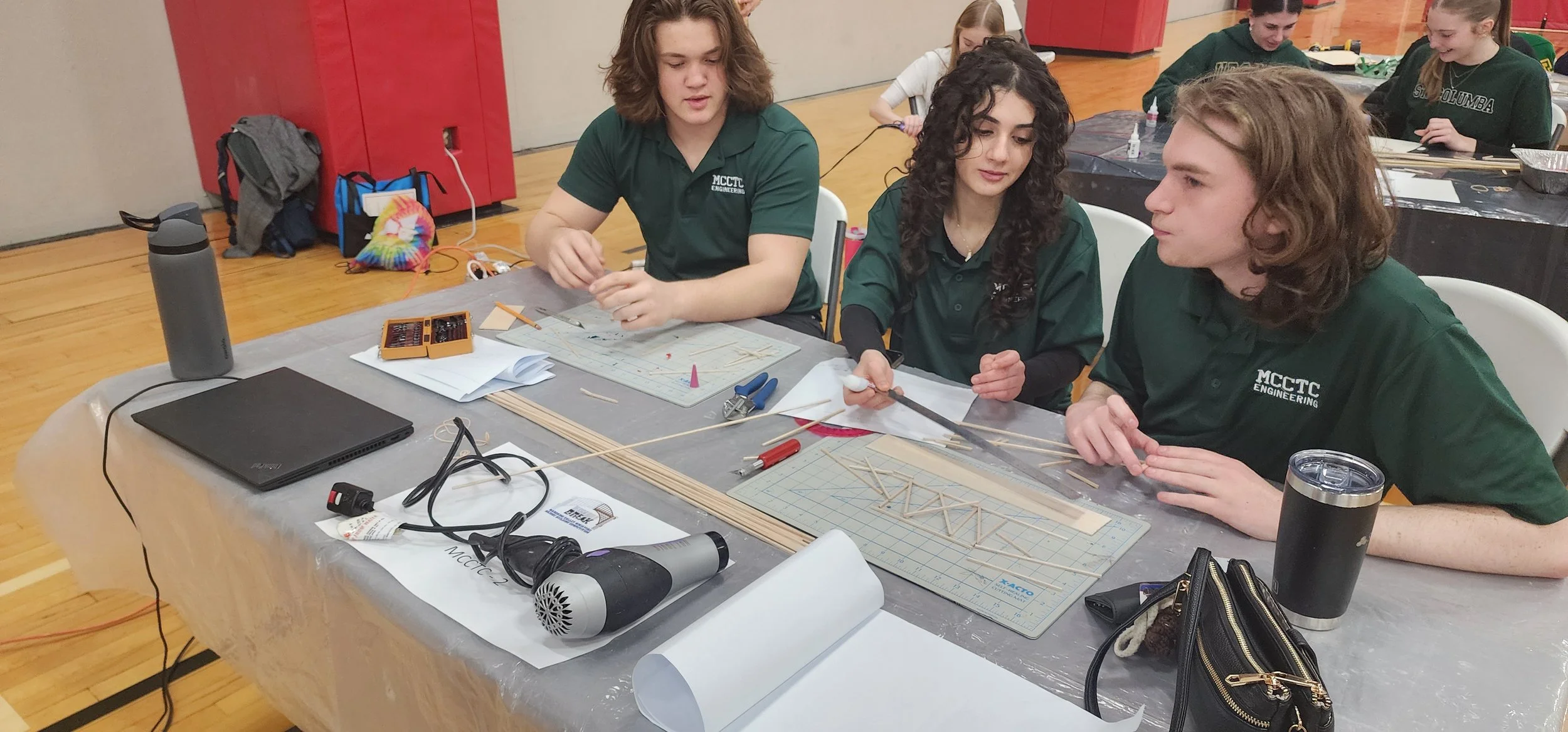 Students in green shirts with 'MCCTC Engineering' logo working on a model bridge at a table with tools and materials in a gymnasium.