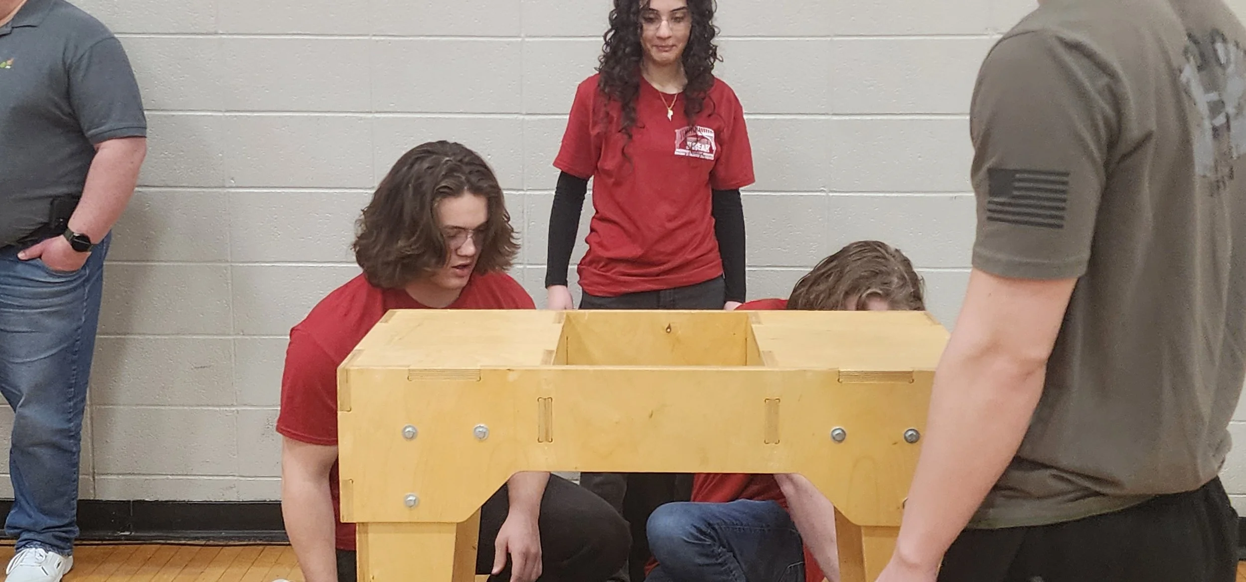 Four people, three women and one man, are gathered around a wooden structure in a room with gray cinder block walls. Two women are kneeling, one looking inside the structure, while the woman standing behind them is observing.