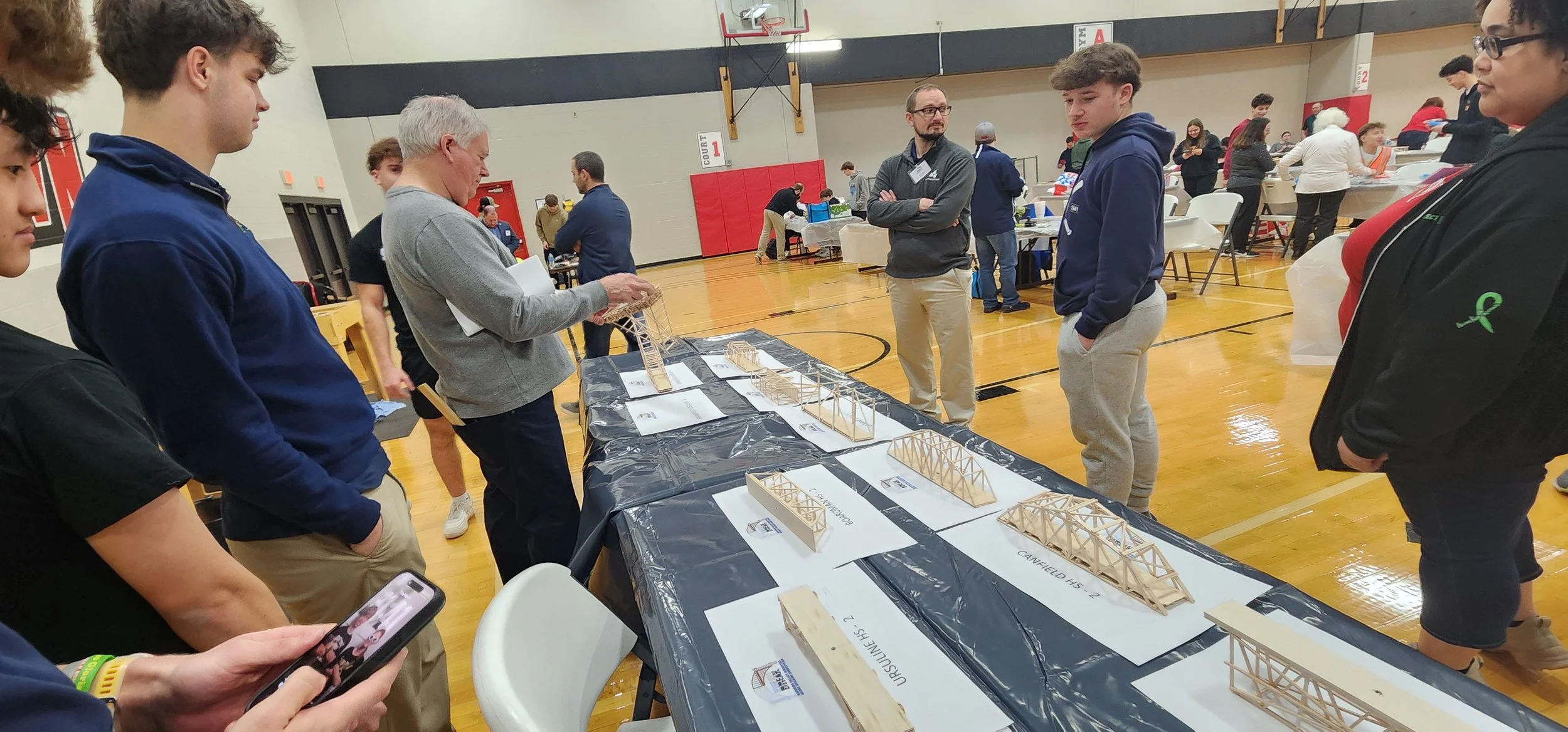 Group of people in a gymnasium looking at wooden bridge models on display tables