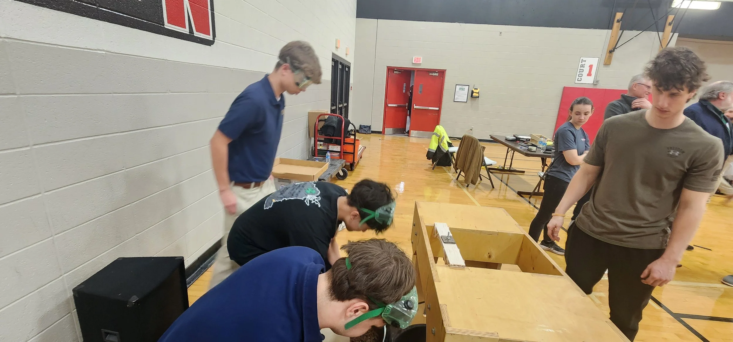 Group of young people wearing safety goggles gathered around a table with a wooden structure in a gymnasium.