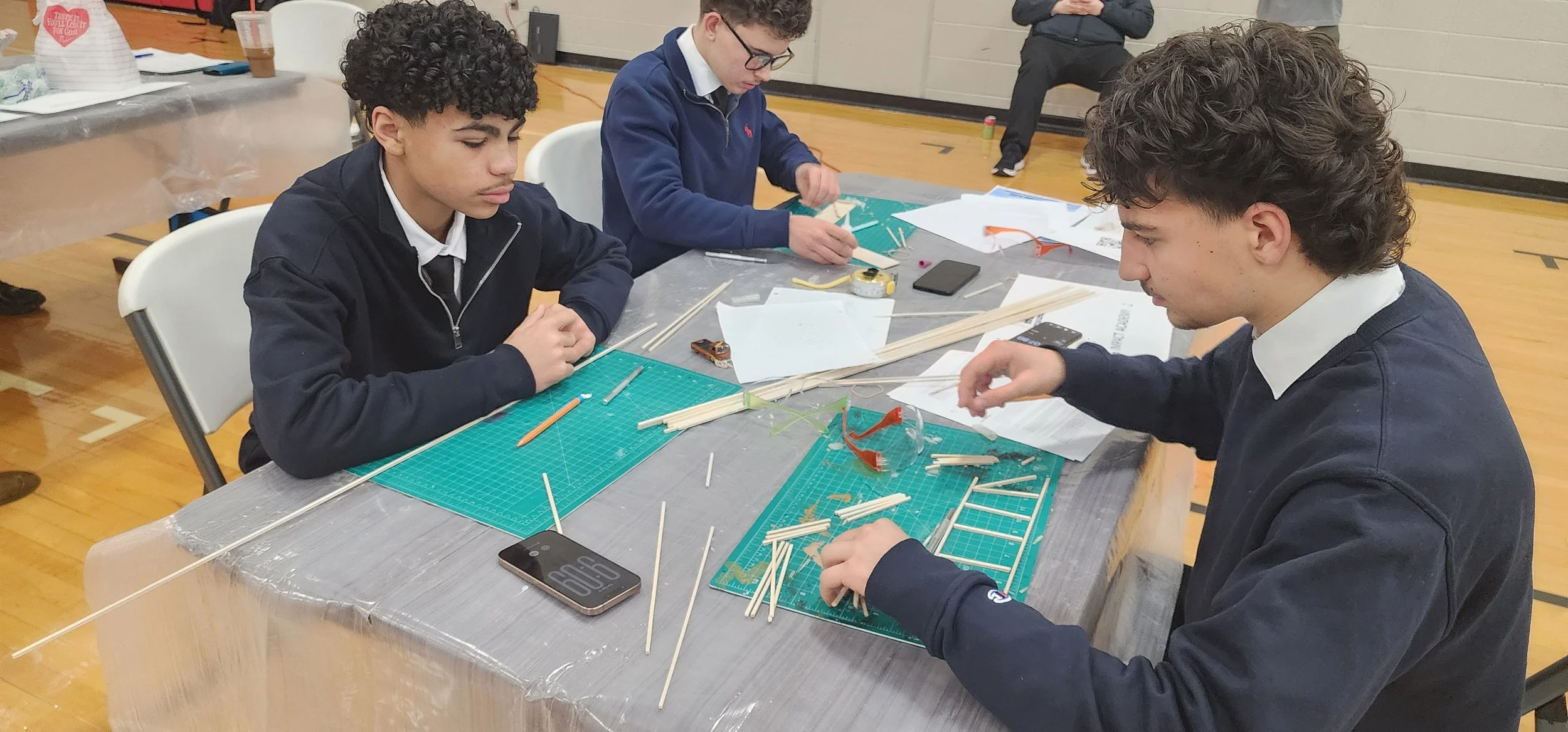 Three young men working on a hands-on STEM or engineering project at a table, assembling a structure with wooden sticks and plastic parts, in a gymnasium or hall.