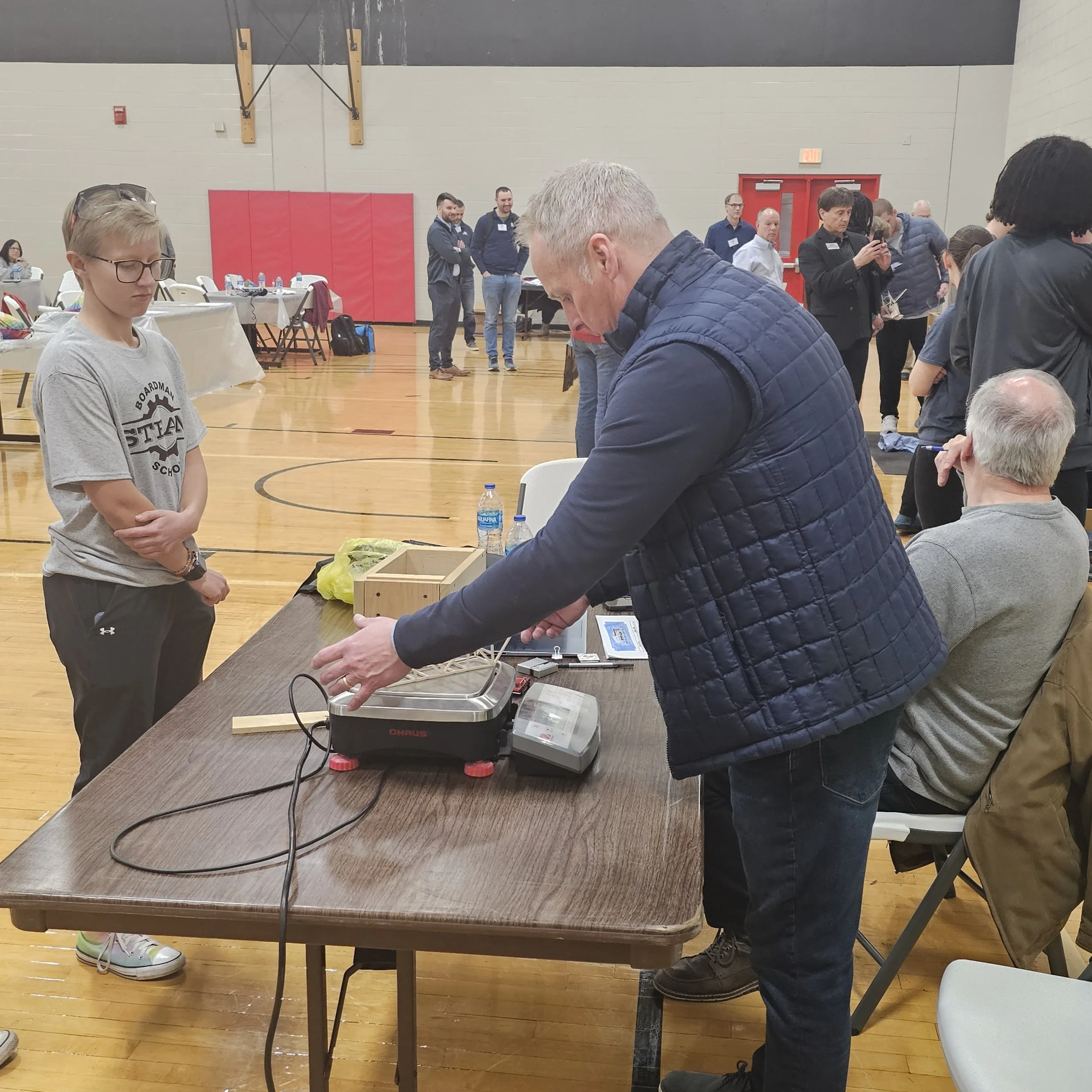 People standing and sitting around tables in a gymnasium during an event. Some individuals are engaged in conversation, and one person is taking a photo with a phone. There are water bottles and a scale on a table in the foreground.