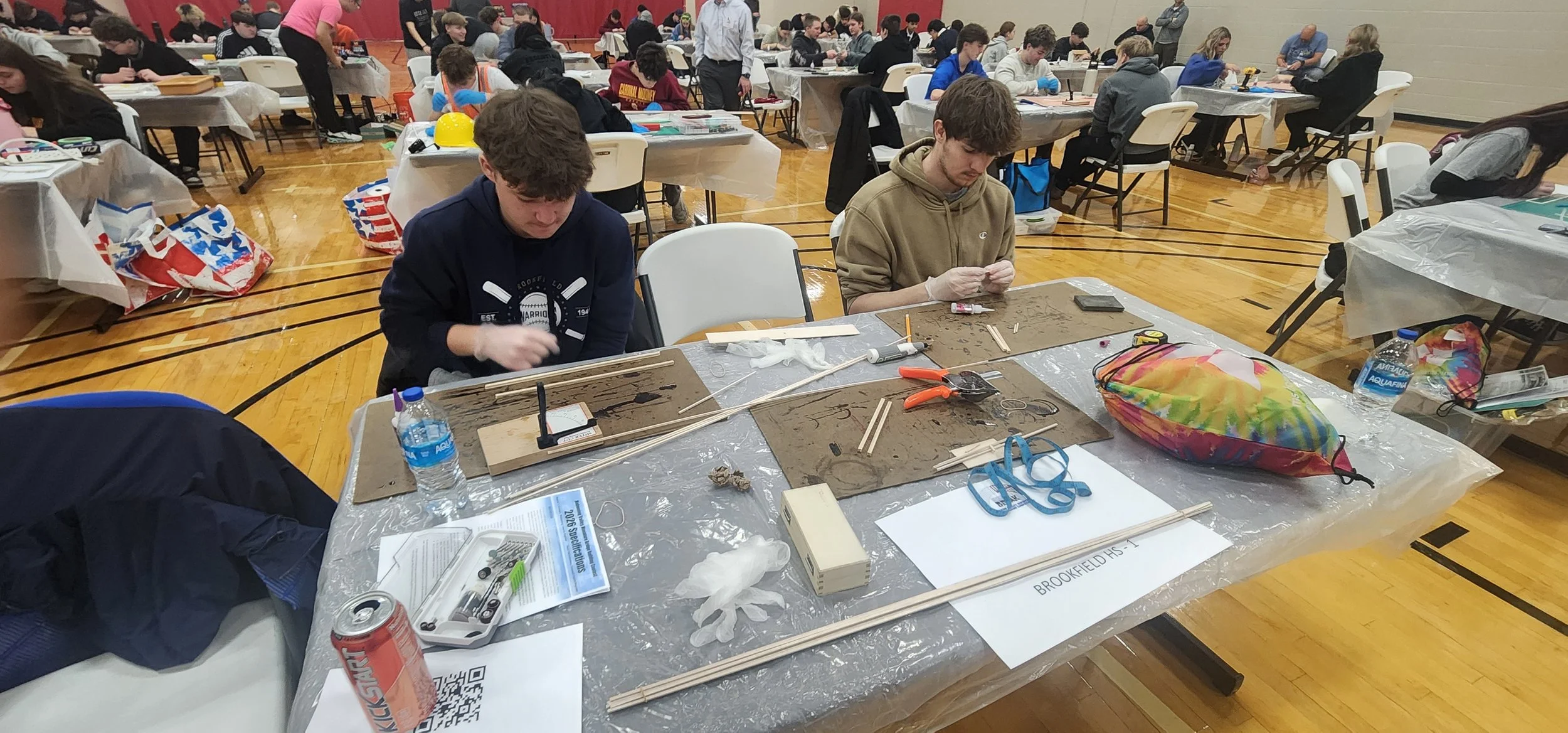 Students working on a STEM project at a large indoor event. They are seated at tables with various materials, tools, and supplies, including wood, tape, scissors, and water bottles. The room is full of people, with some standing and observing.