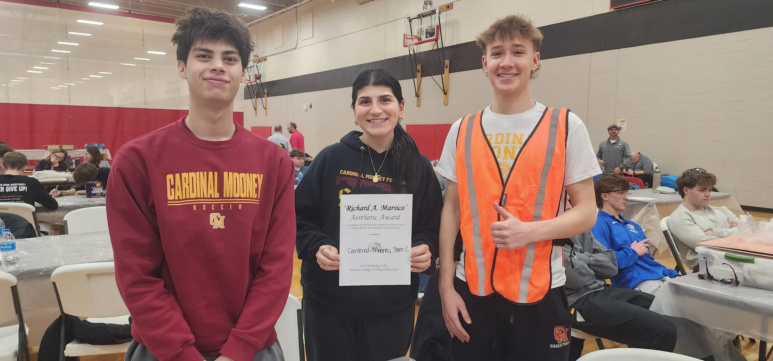 Three young people standing together indoors, smiling. The person in the middle is holding a certificate that reads 'Richard A. Marsico Aesthetic Award' and is wearing a black hoodie with yellow lettering and a gold necklace. The individual on the le