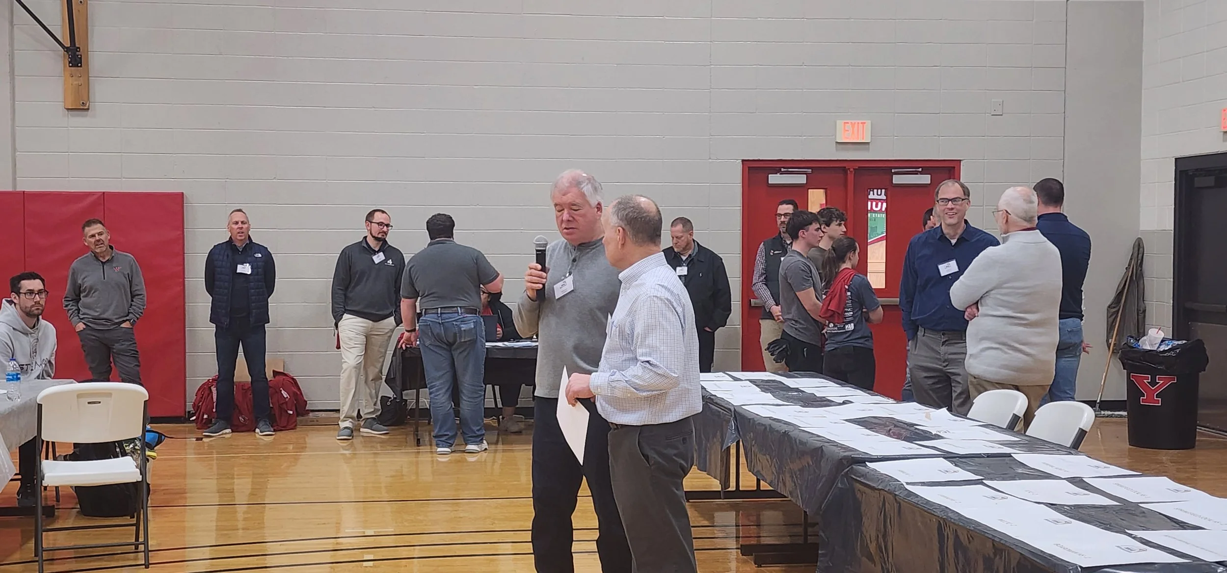 People gathered in a gymnasium for an event, with some speaking and others listening, tables with documents, and a mix of seated and standing attendees.
