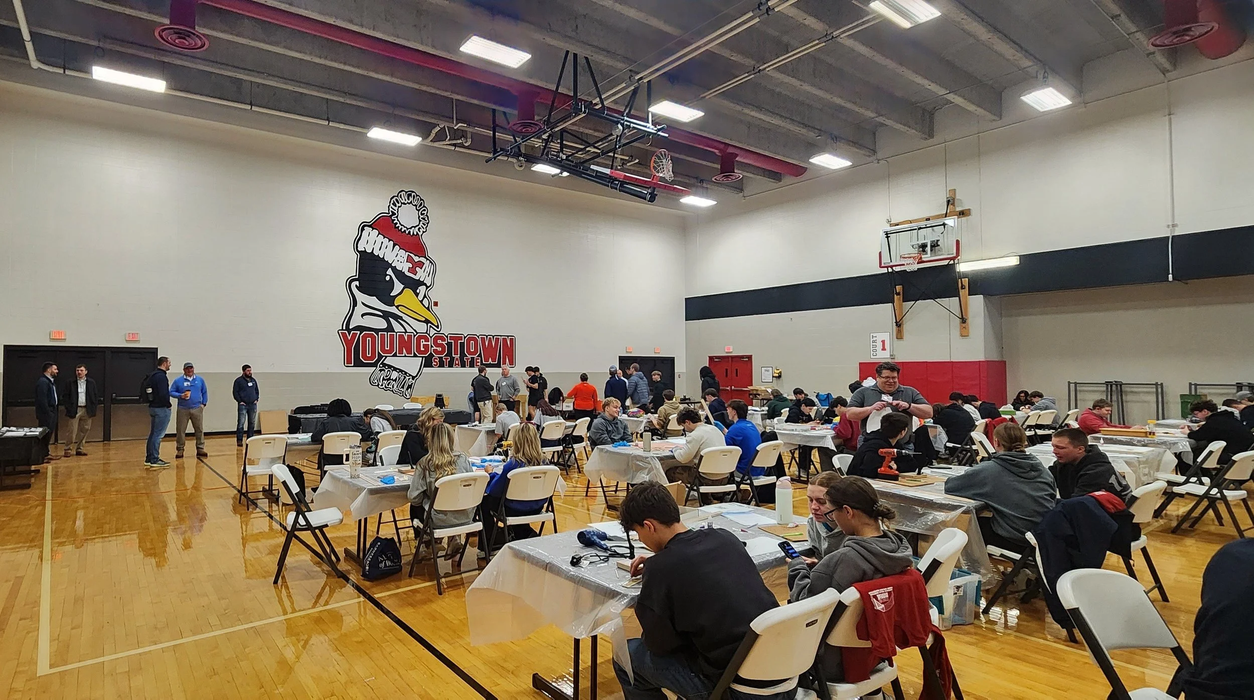 An indoor event with people sitting at tables in a gymnasium, some working with papers and tools. There is a large YouthTown State logo with a cartoon duck on the wall, and several individuals standing and walking around.
